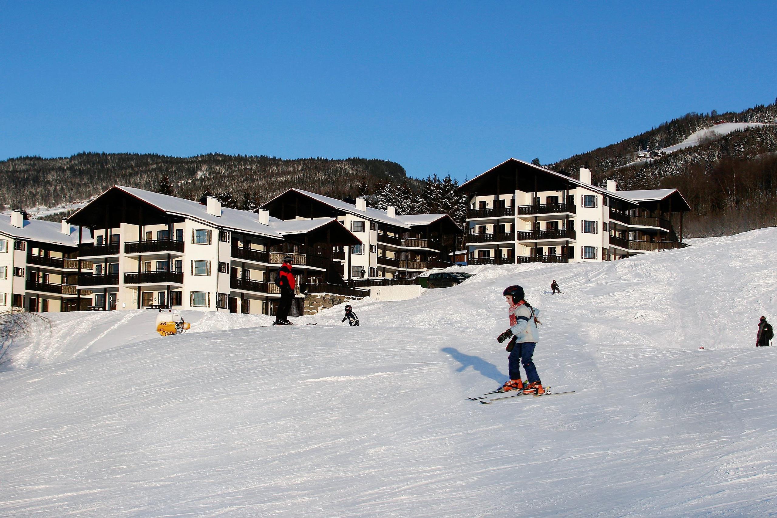 Barn står på ski i en solfylt skibakke med leilighetsbygg, grantrær og snødekte fjell under blå himmel..