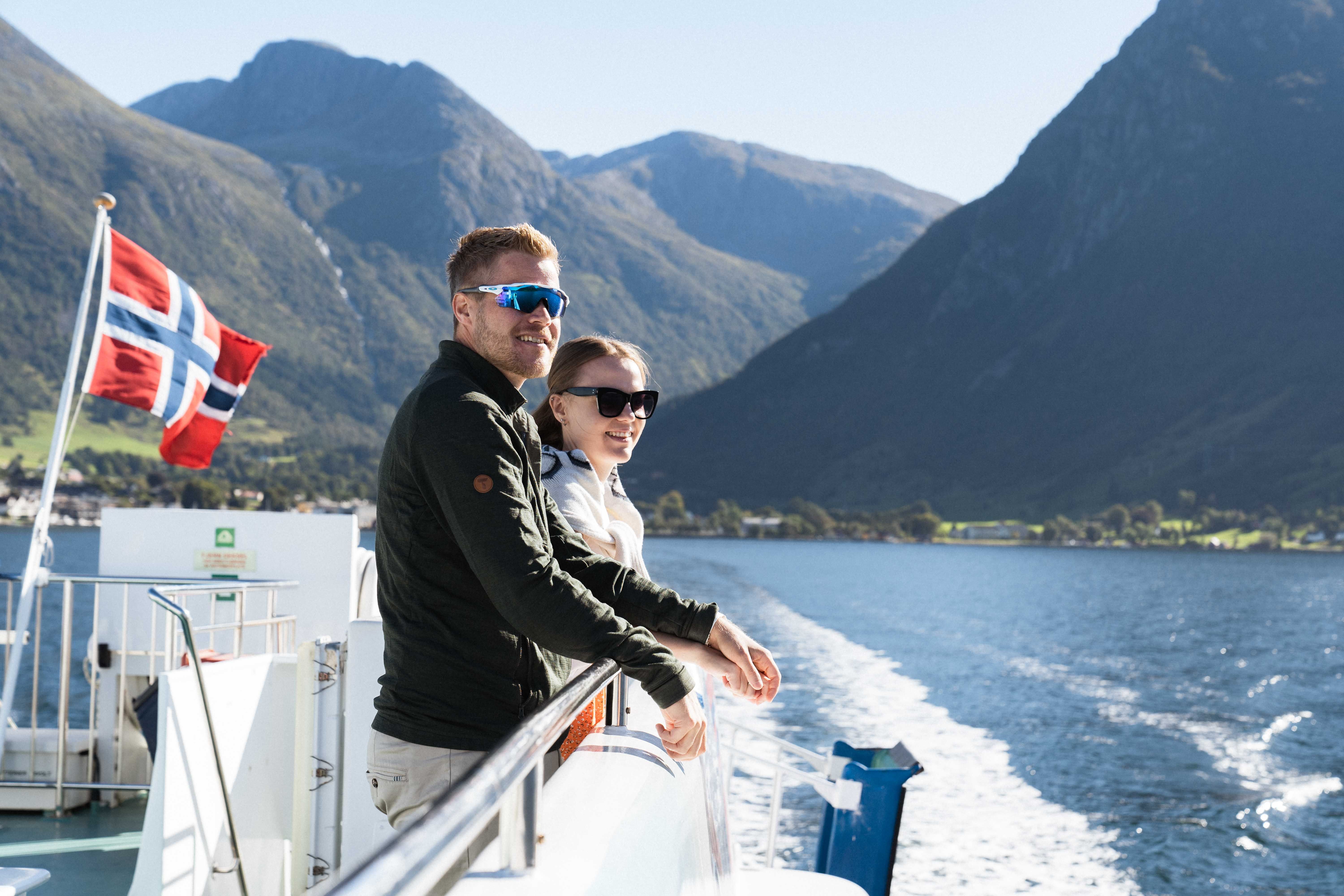 A smiling couple on Hardangerfjordekspressen with the Norwegian flag and scenic surroundings in the background.
