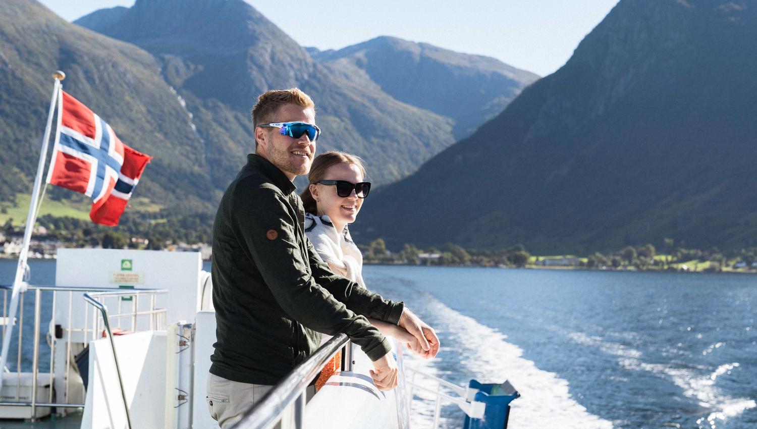 A smiling couple on Hardangerfjordekspressen with the Norwegian flag and scenic surroundings in the background.