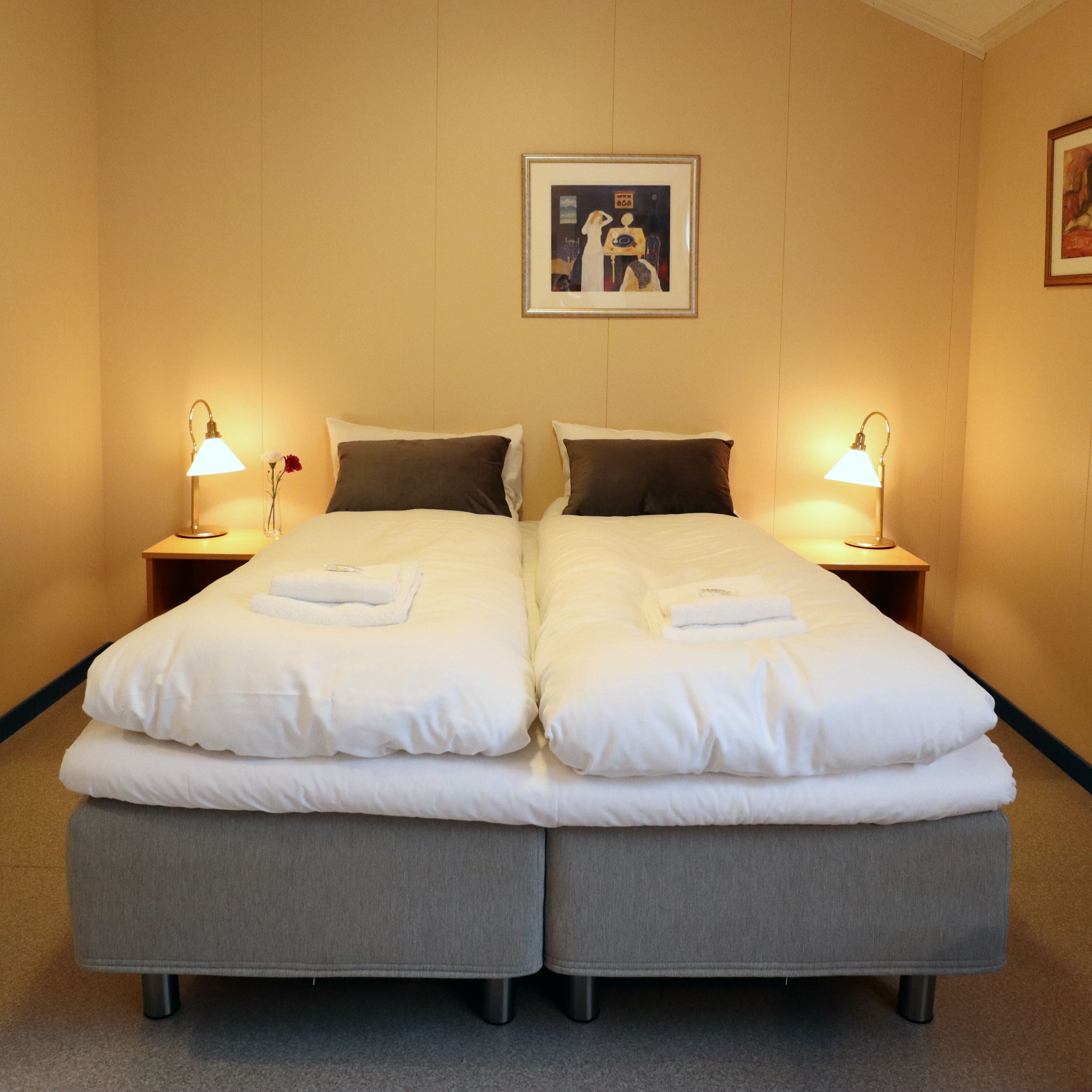 Two twin beds with white duvets, folded towels, bedside lamps, and framed art on a beige wall..