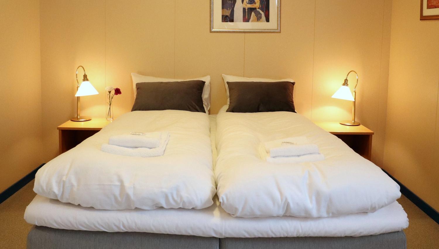 Two twin beds with white duvets, folded towels, bedside lamps, and framed art on a beige wall..