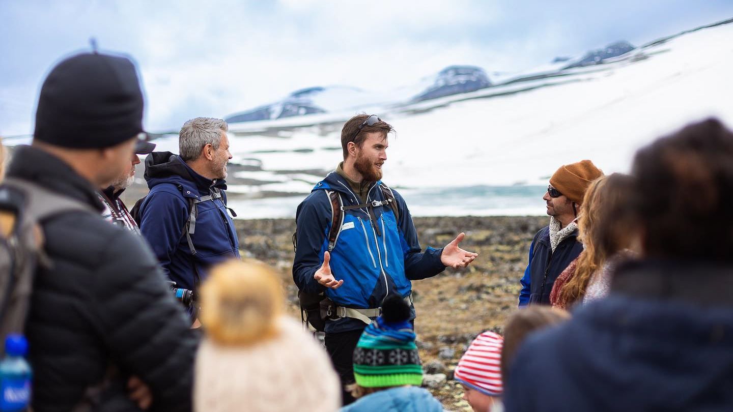 Ein guide i blå jakke snakkar til ei gruppe ute på steinete mark, med snødekte fjell i bakgrunnen..