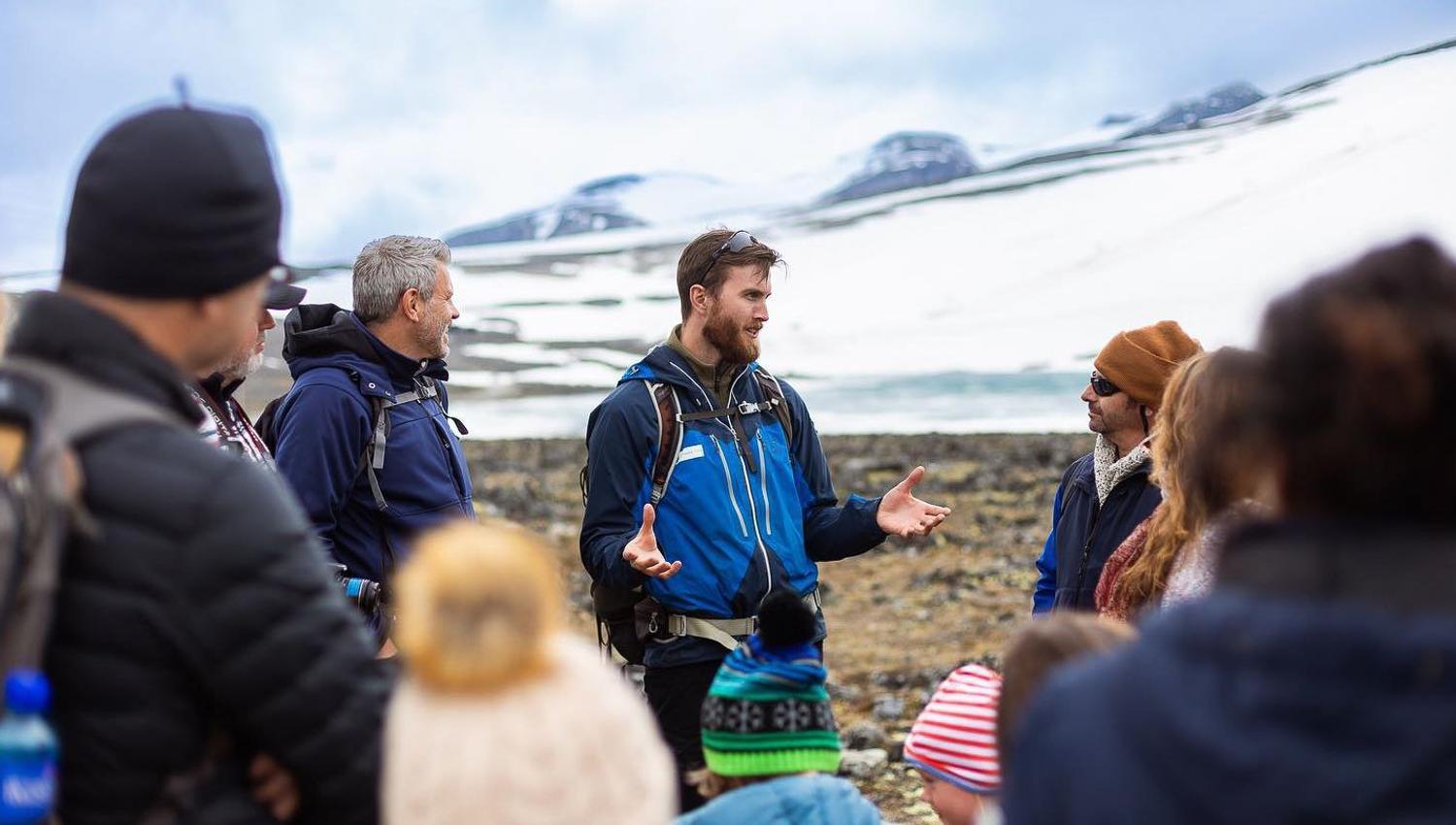 Ein guide i blå jakke snakkar til ei gruppe ute på steinete mark, med snødekte fjell i bakgrunnen..