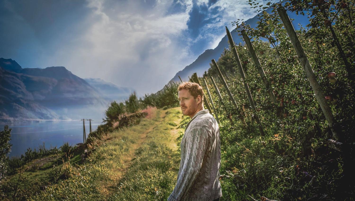 Man walking through the orchards of Hardangerfjord with the fjord in the background.