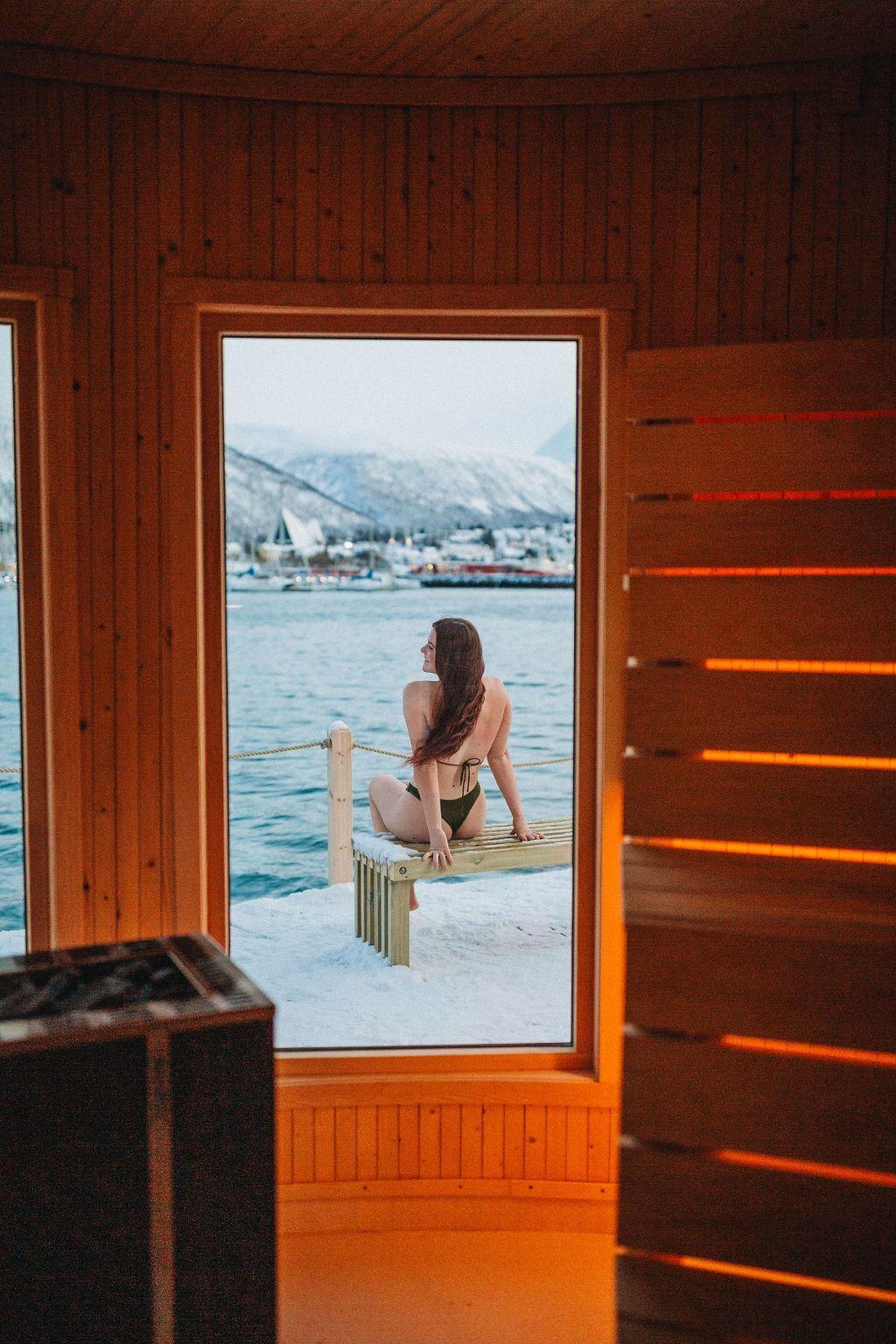 A guest sitting by the open sauna doorway with views across the water and surrounding mountains in Tromsø.