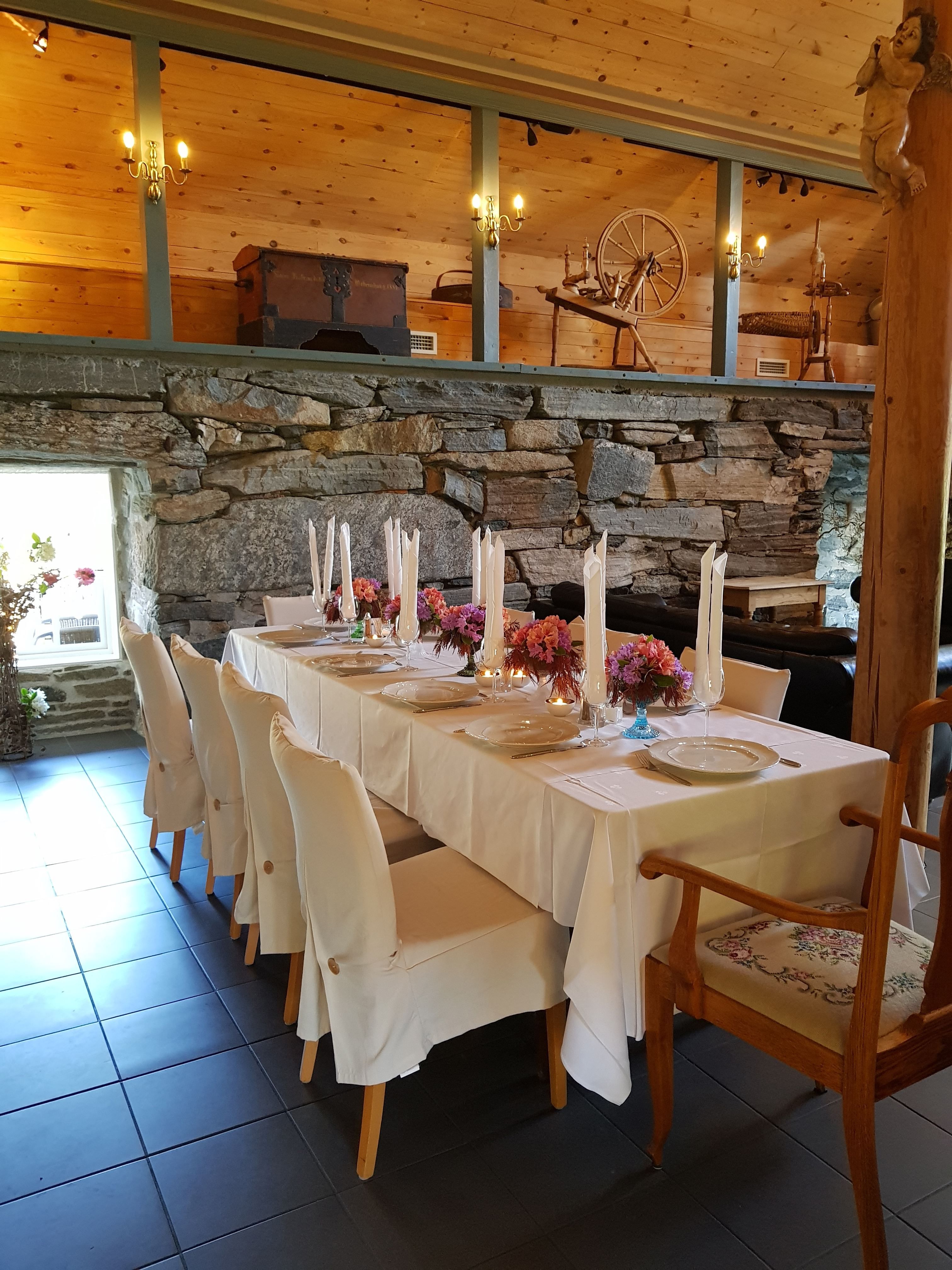 Orheimstunet in Nordfjord - Long white-clothed dining table with candles and flowers in a rustic room with stone wall and wooden loft decor..