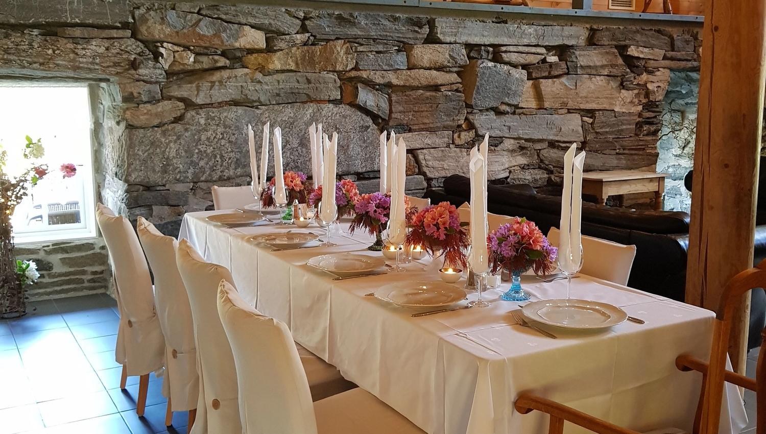 Orheimstunet in Nordfjord - Long white-clothed dining table with candles and flowers in a rustic room with stone wall and wooden loft decor..