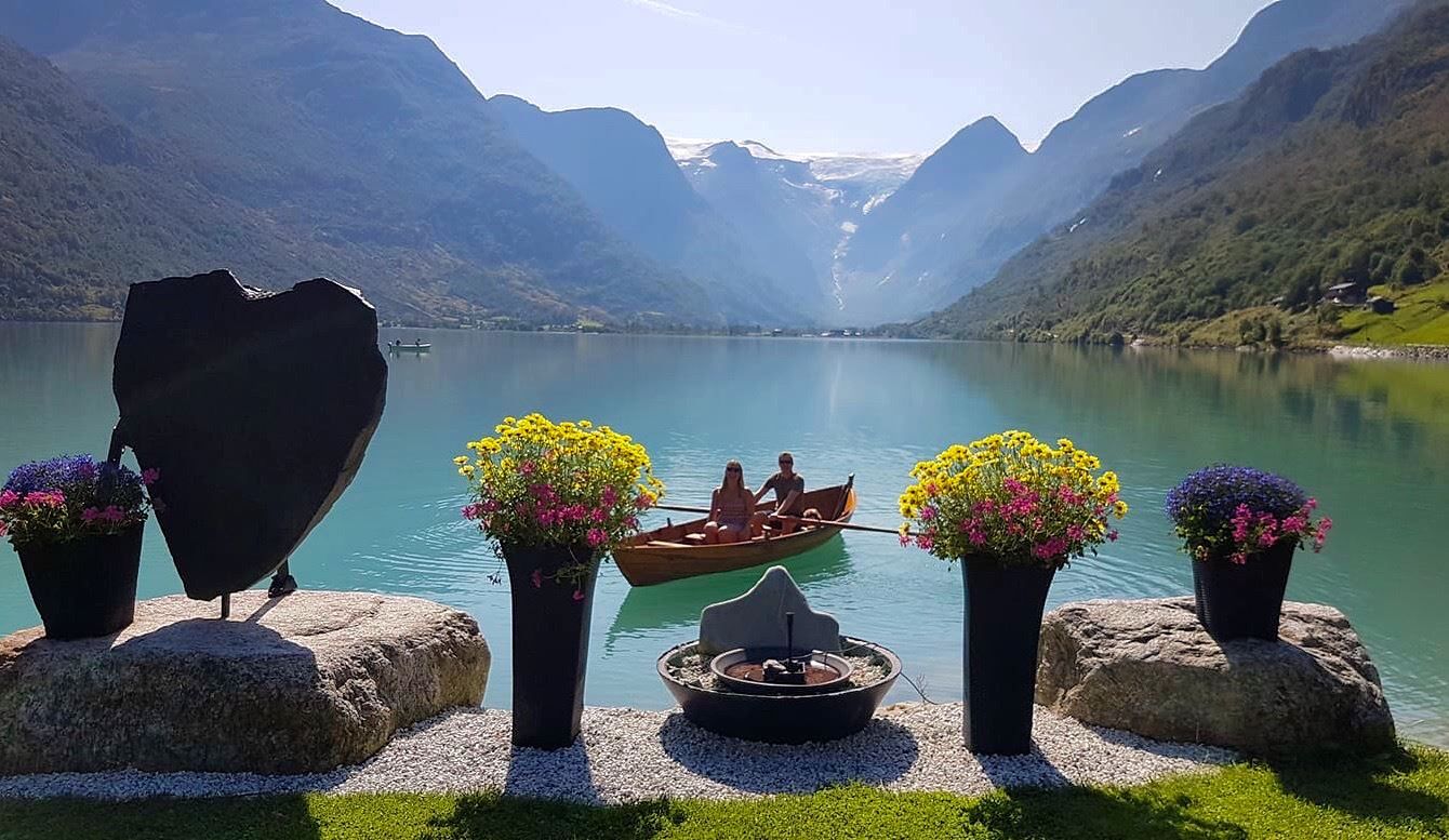 Two people sit in a small rowboat on a turquoise lake, framed by potted flowers and steep mountains in the background.