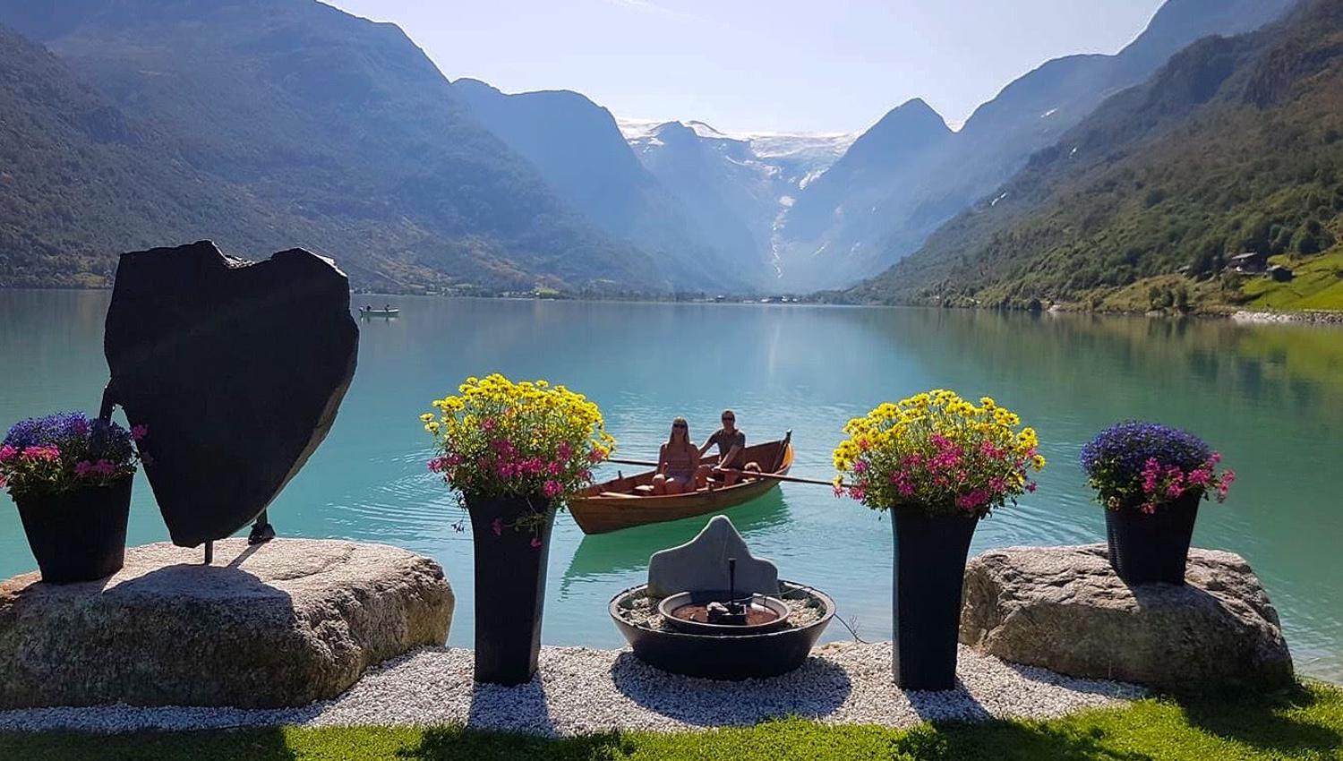 Two people sit in a small rowboat on a turquoise lake, framed by potted flowers and steep mountains in the background.