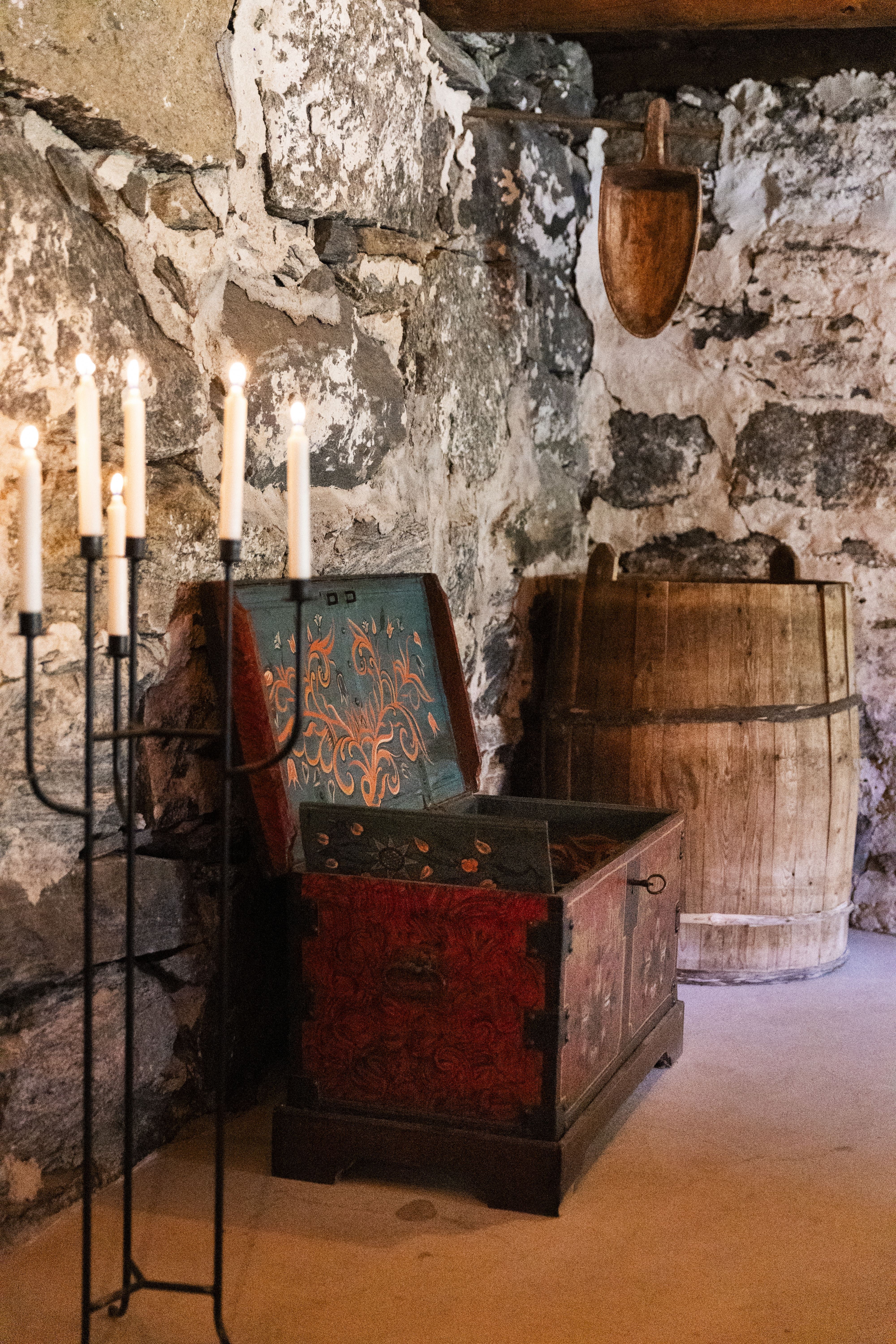 Open painted wooden chest beside a barrel and candle stand in a rustic stone-walled room with wooden beams.