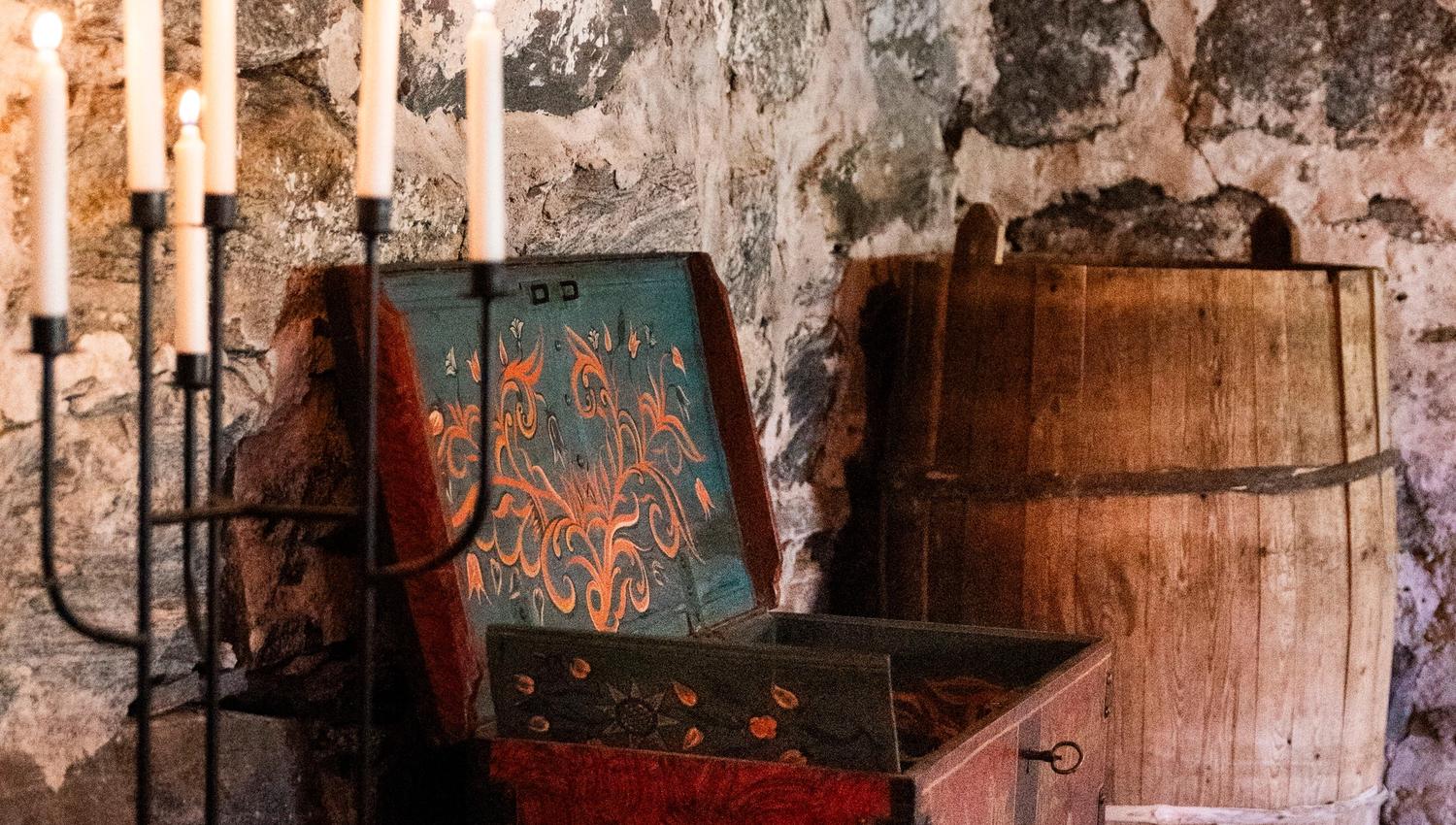 Open painted wooden chest beside a barrel and candle stand in a rustic stone-walled room with wooden beams.