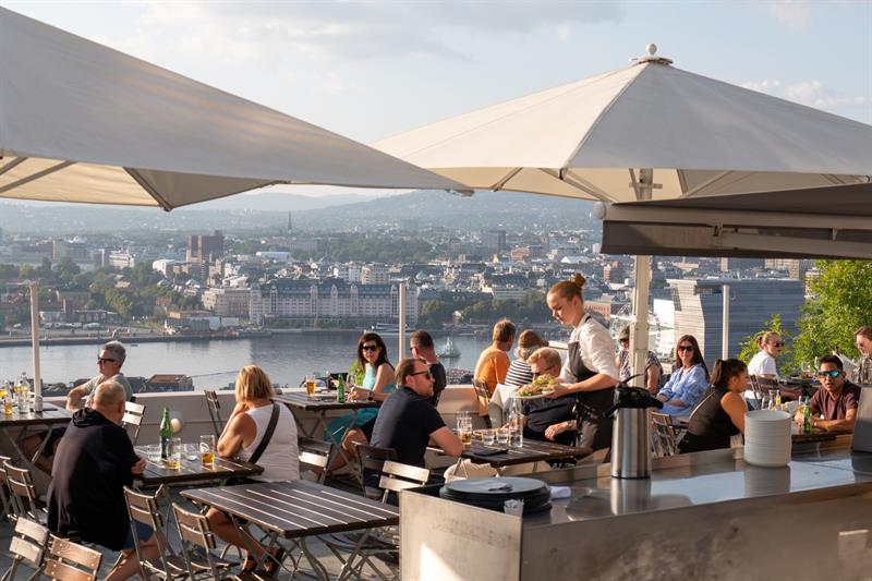 Servitør serverer mat til gjester på en travel takterrasse med utsikt over byen.
