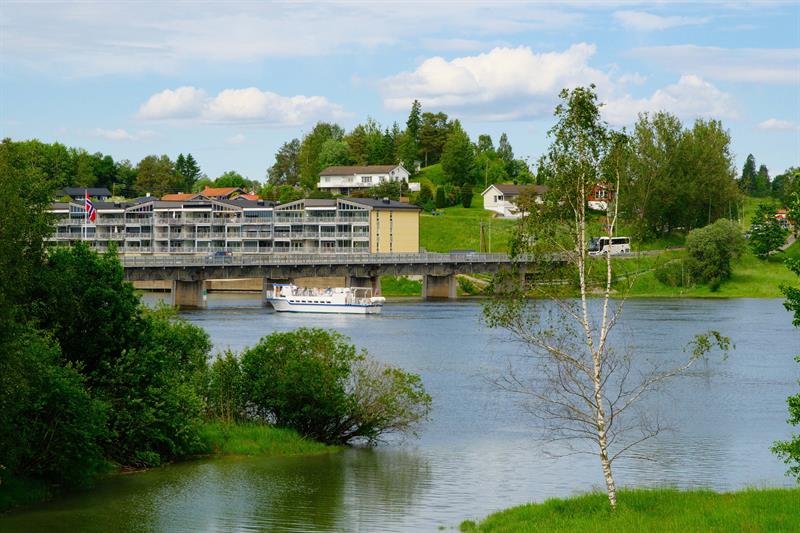 Rolig elv omgitt av grønt landskap, med flere bygninger langs vannkanten i bakgrunnen og en båt som seiler under en bro