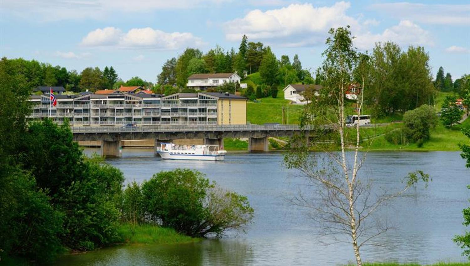 Rolig elv omgitt av grønt landskap, med flere bygninger langs vannkanten i bakgrunnen og en båt som seiler under en bro