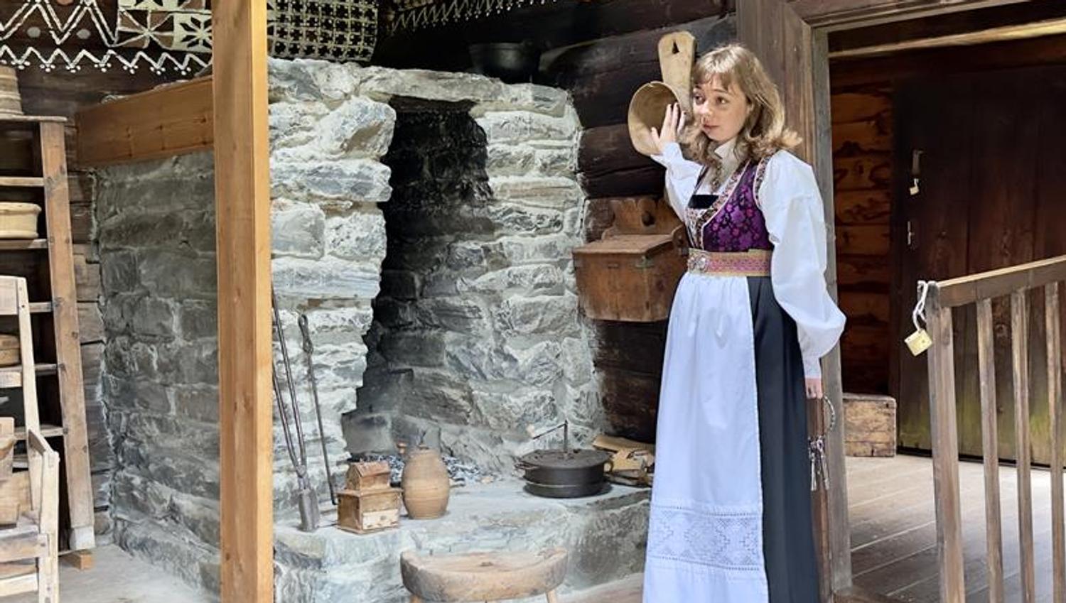 i kvinne i Hardangerbunad har omvising i ein av stovene på Hardanger folkemuseum. Ho står foran et ildsted av stein inne i en gammel lafta stove, det er dekor på treveggene og gamle gjenstander står på gulvet og henger på veggen