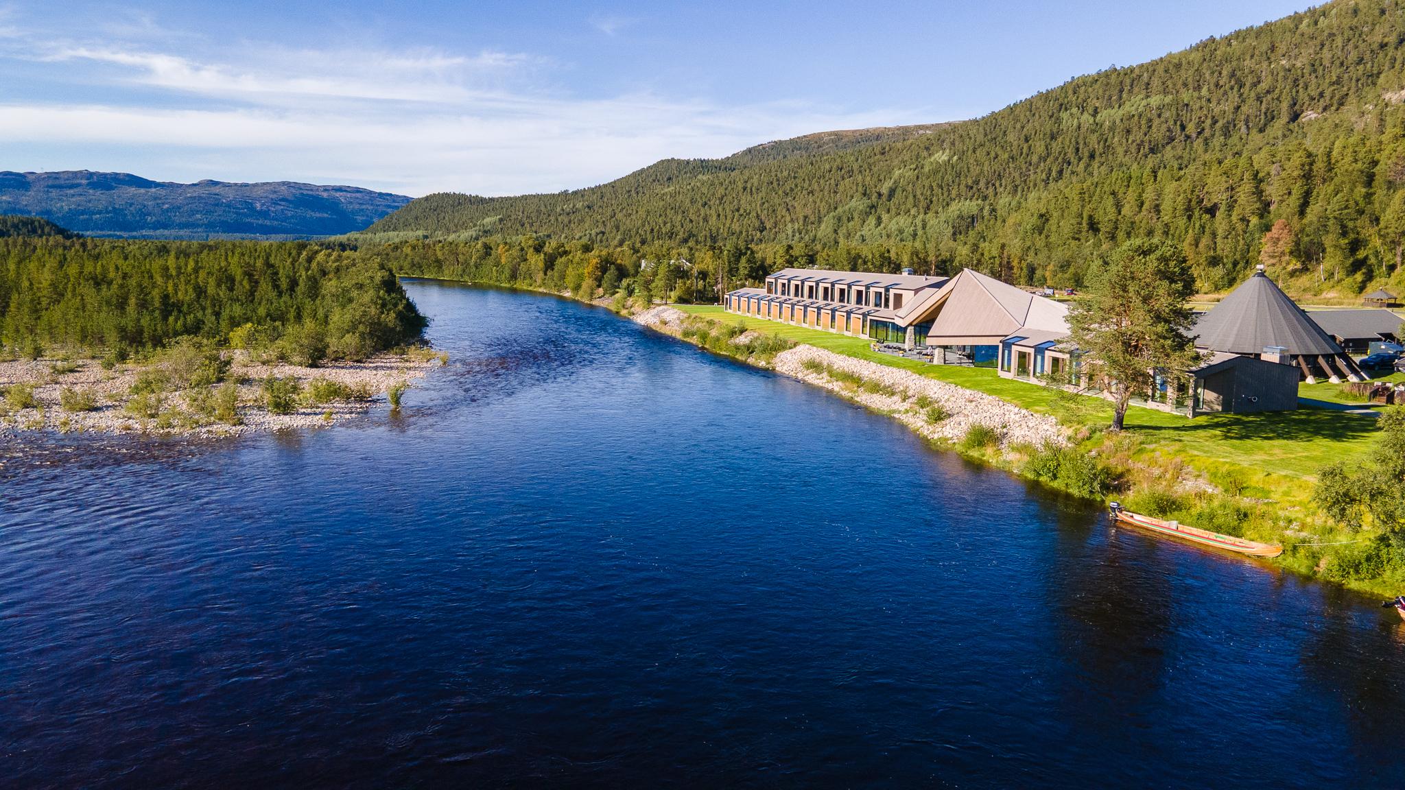 Vue aérienne d'un hôtel moderne au bord d'une rivière entourée de forêts verdoyantes.