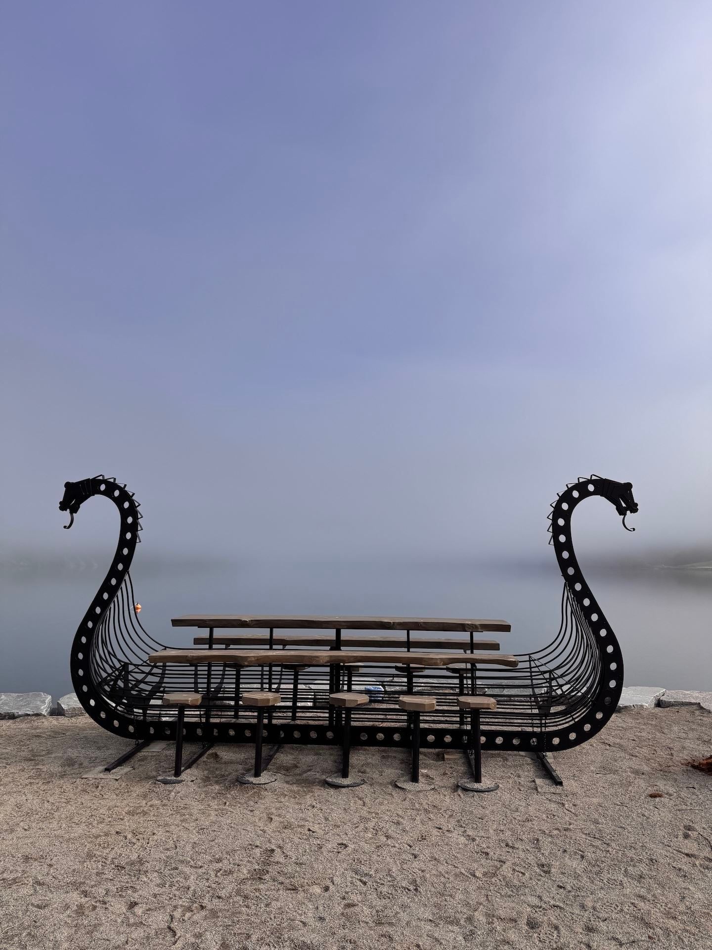 Metal Viking ship-shaped bench with wooden seats on a rocky shore, facing calm water under a hazy sky..
