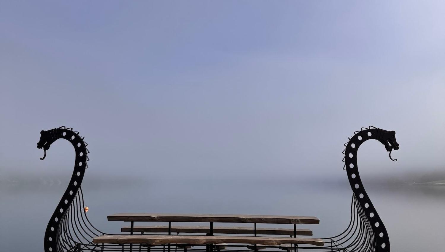 Metal Viking ship-shaped bench with wooden seats on a rocky shore, facing calm water under a hazy sky..