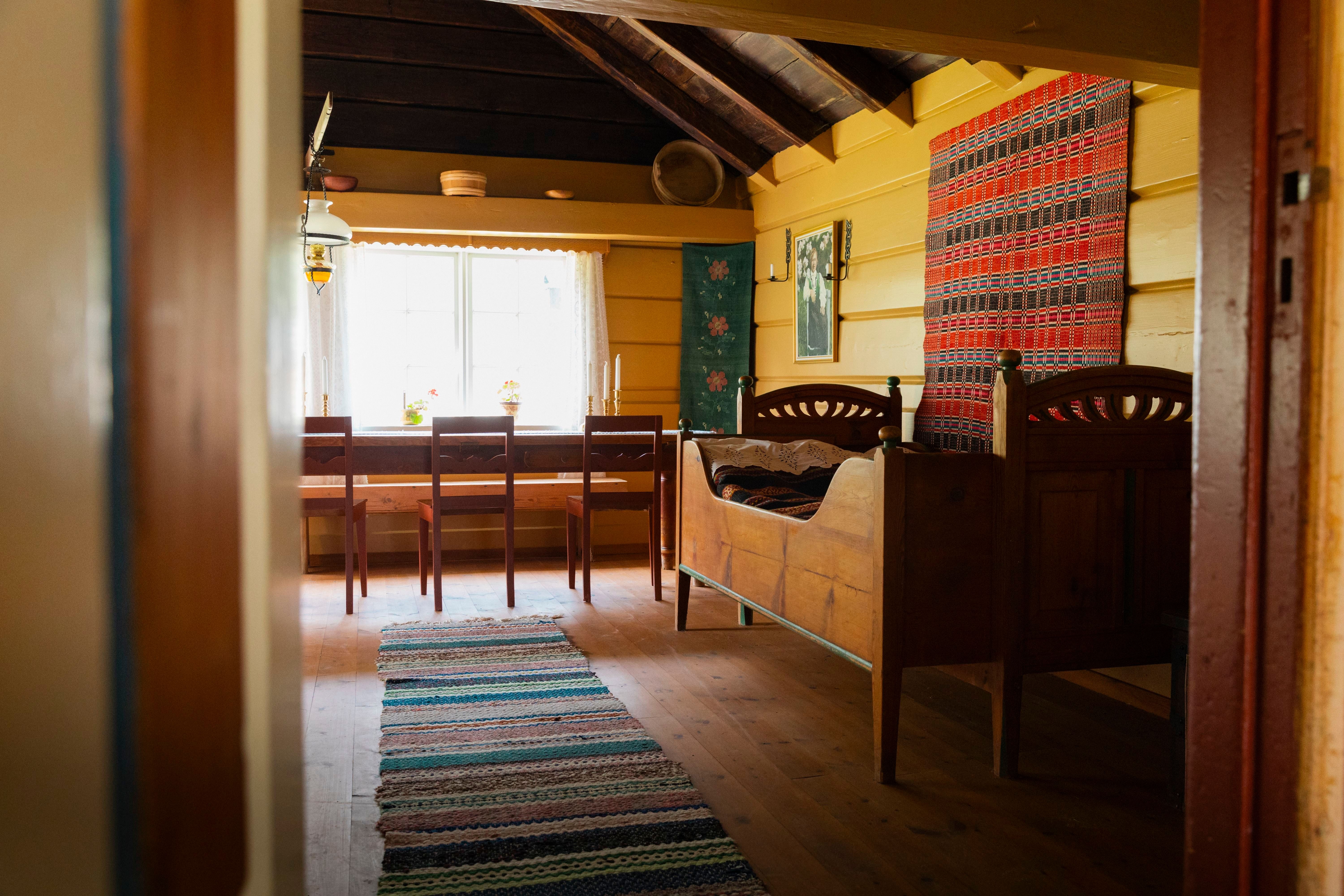 Sunlit rustic cabin room with wooden bed, striped wall tapestry, dining table by a window, and woven rug on floor.