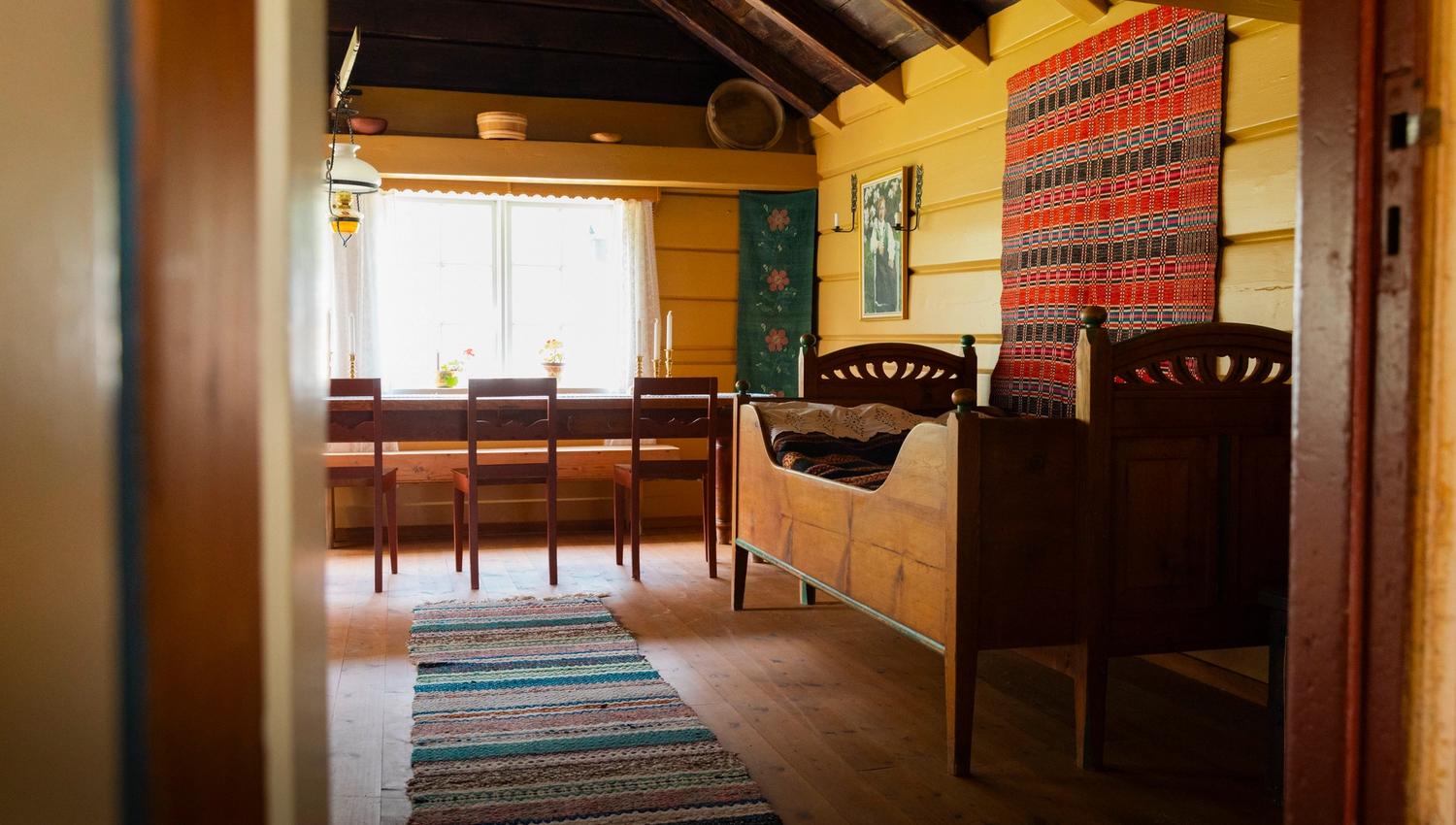 Sunlit rustic cabin room with wooden bed, striped wall tapestry, dining table by a window, and woven rug on floor.
