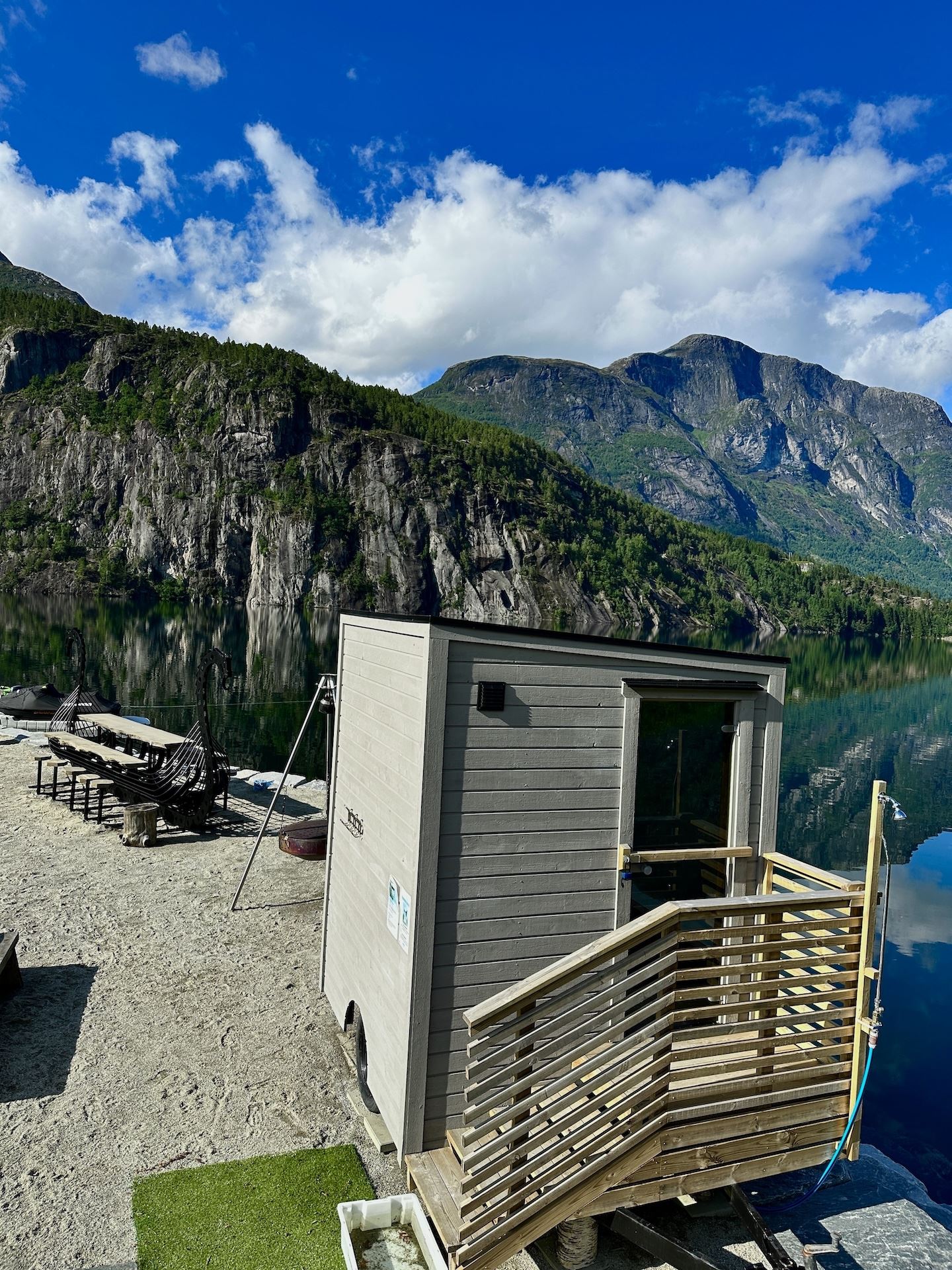 Small gray sauna cabin on a lakeshore with lounge chairs, calm water reflections, and rocky green mountains under clouds..