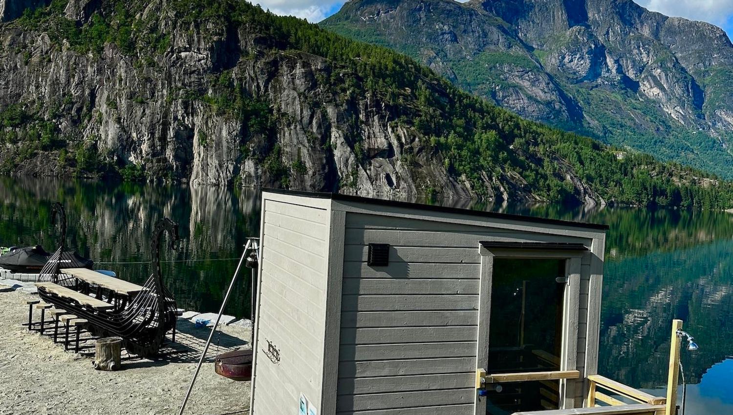 Small gray sauna cabin on a lakeshore with lounge chairs, calm water reflections, and rocky green mountains under clouds..