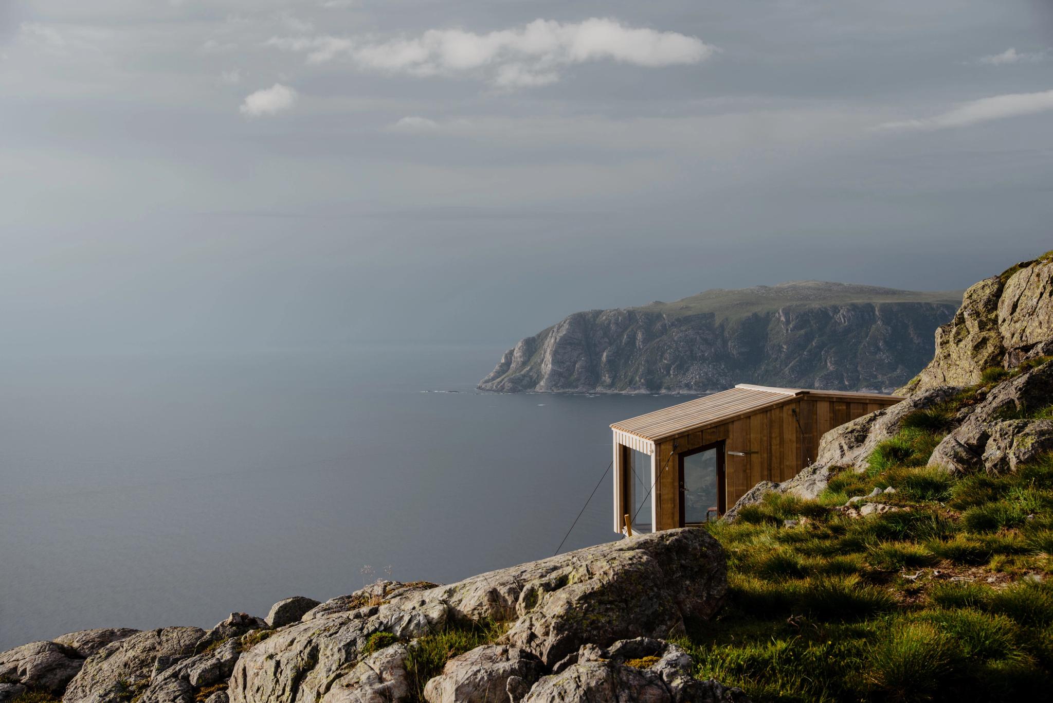 A small wooden cabin sits on a rocky, grassy cliff overlooking a calm ocean with distant headlands under clouds..