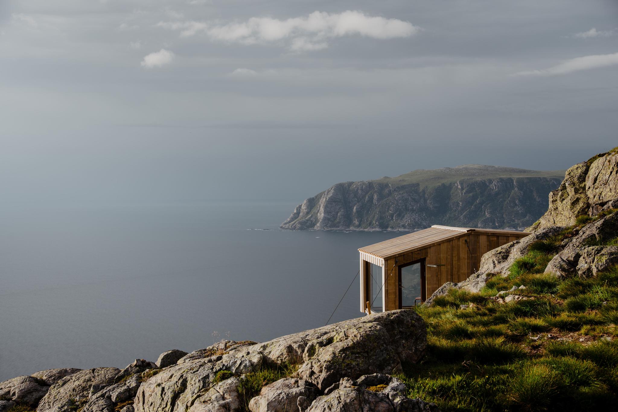 A small wooden cabin sits on a rocky, grassy cliff overlooking a calm ocean with distant headlands under clouds..