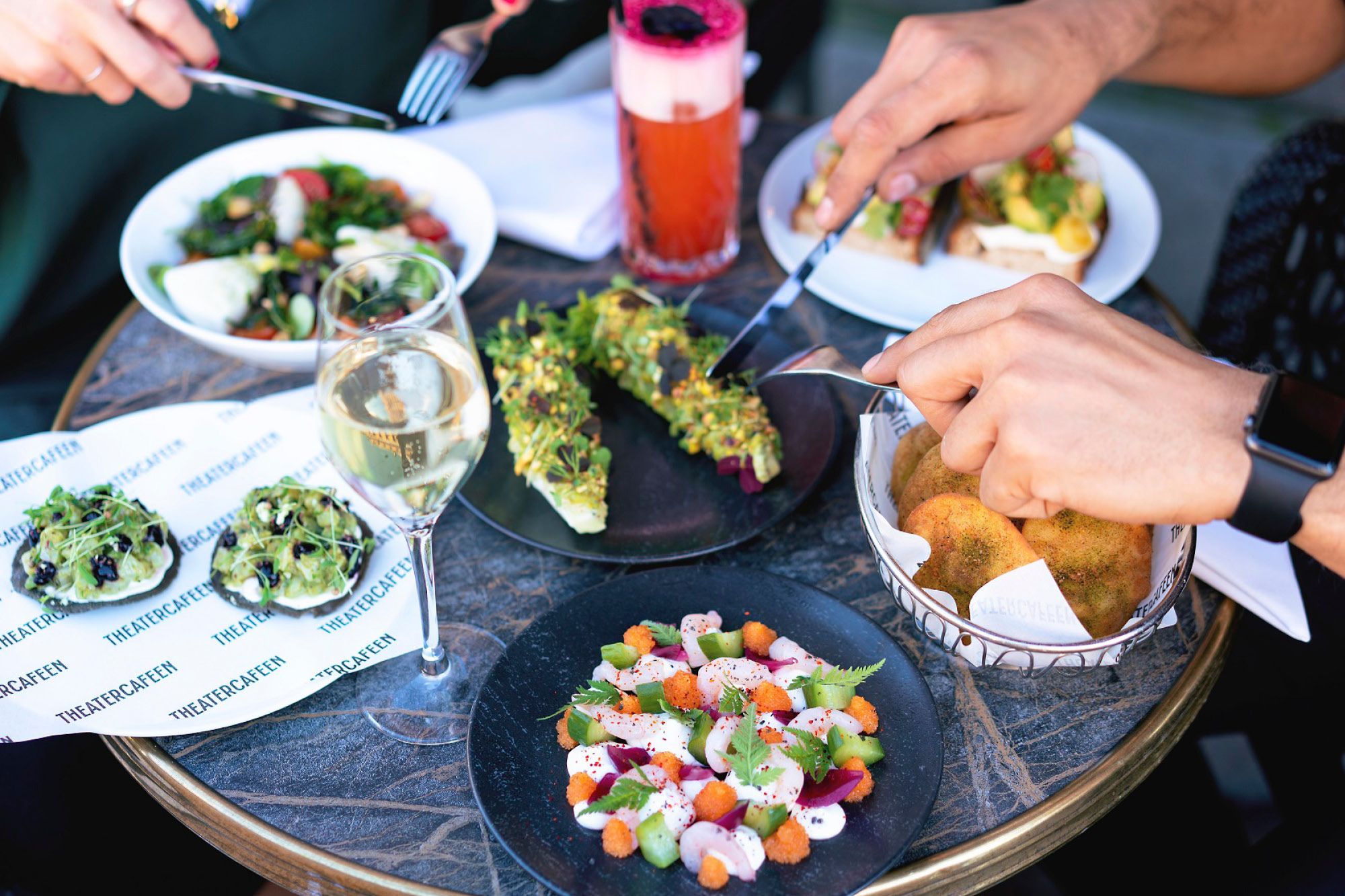 Various dishes, a glass of white wine and a drink on a table, four hands cutting the food with cutlery.