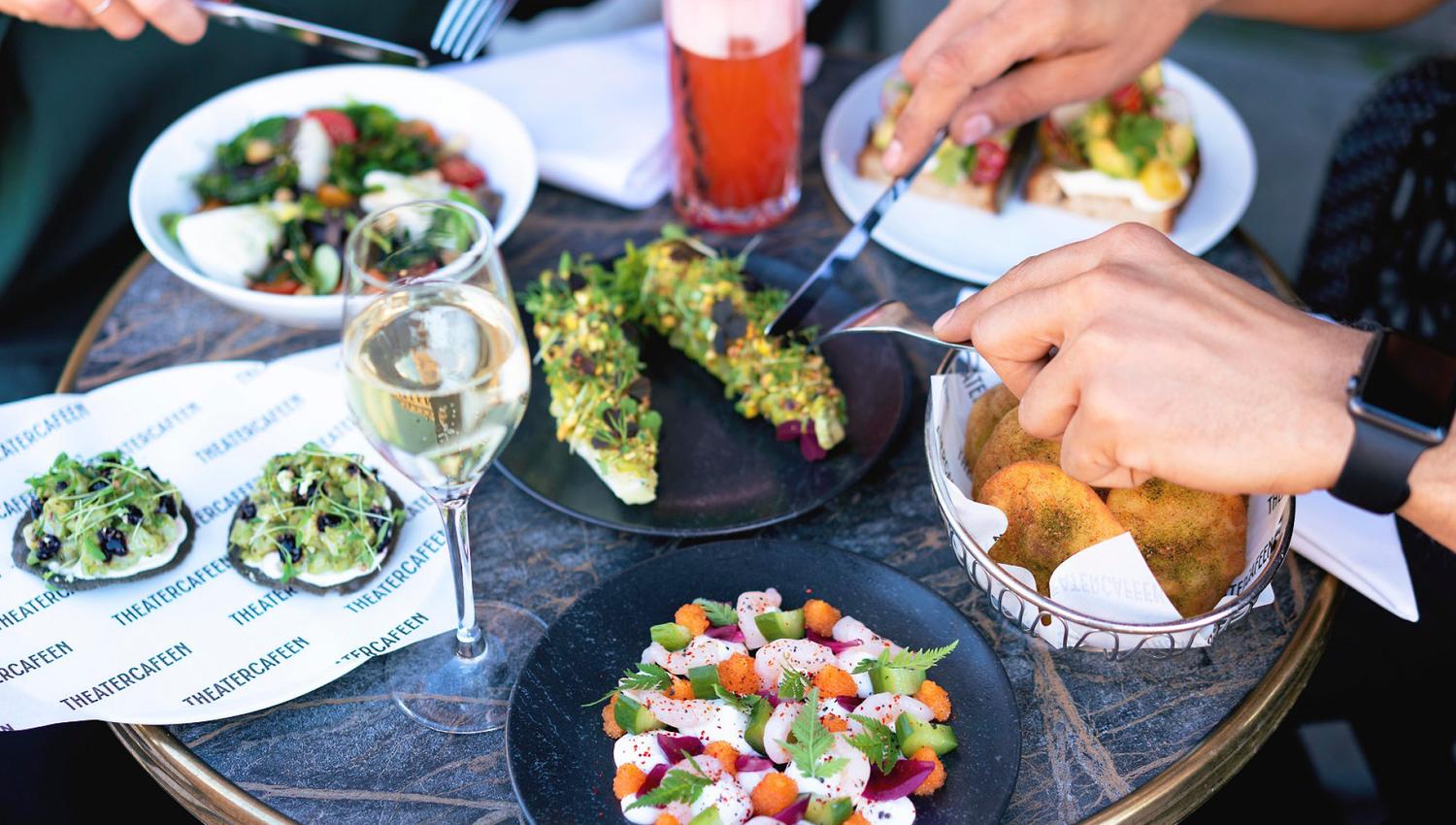 Various dishes, a glass of white wine and a drink on a table, four hands cutting the food with cutlery.