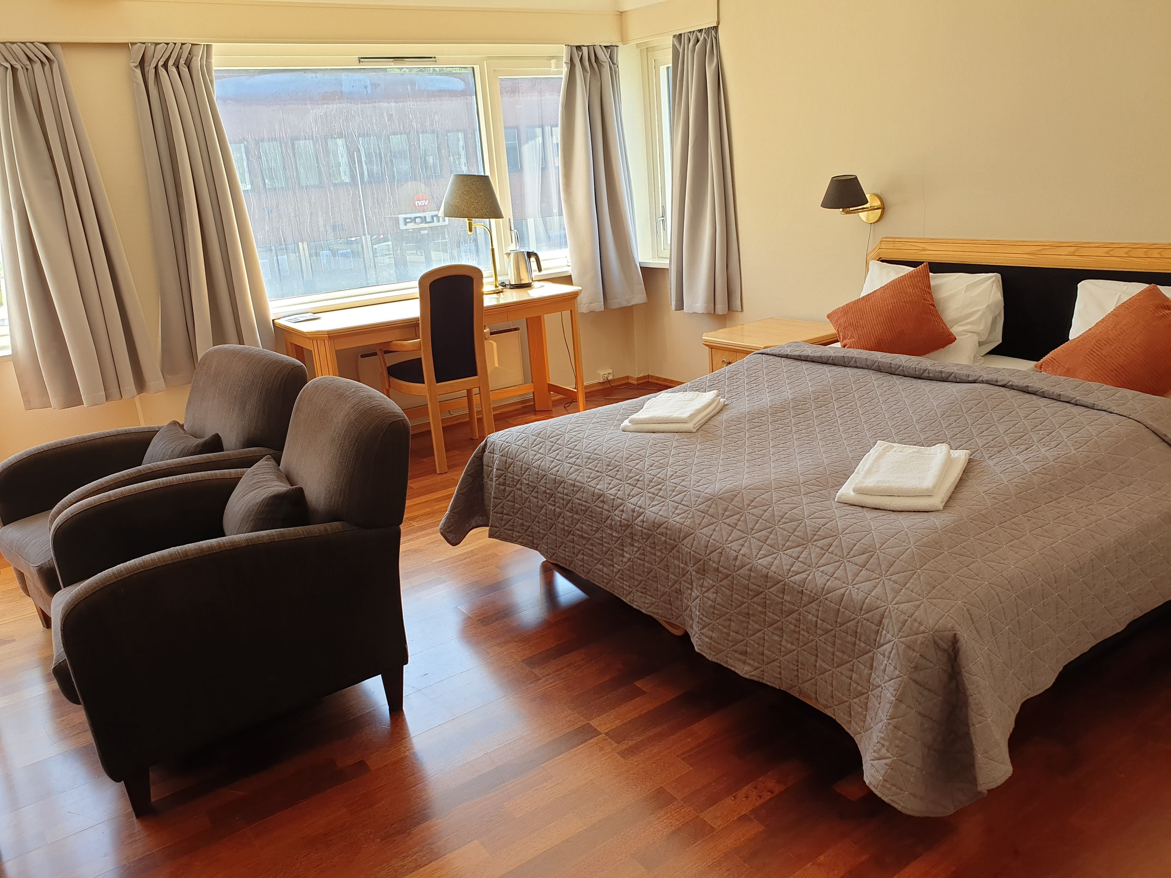 Eit ryddig hotellrom med dobbeltseng, grått sengeteppe og handkle, skrivebord ved vindauge og to stolar..