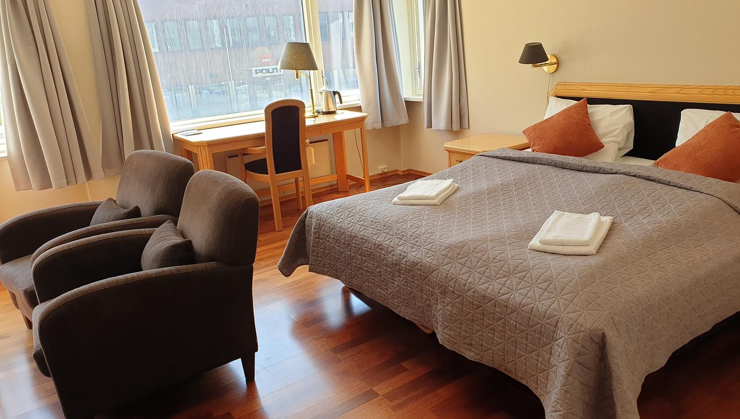 Eit ryddig hotellrom med dobbeltseng, grått sengeteppe og handkle, skrivebord ved vindauge og to stolar..
