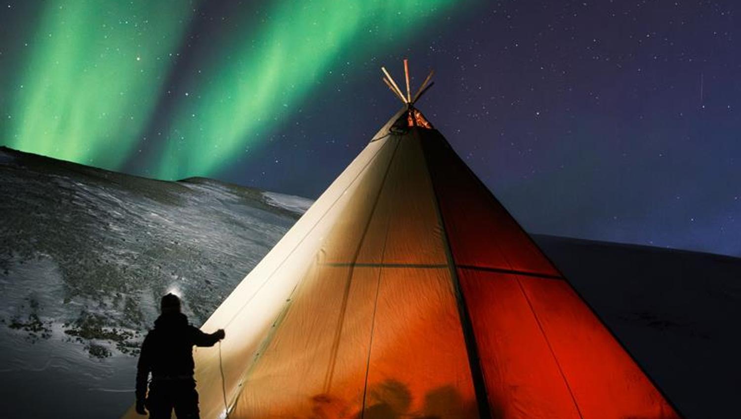 A lighted tent. A person standing outside with headlamp. Green northern ligths on the sky.