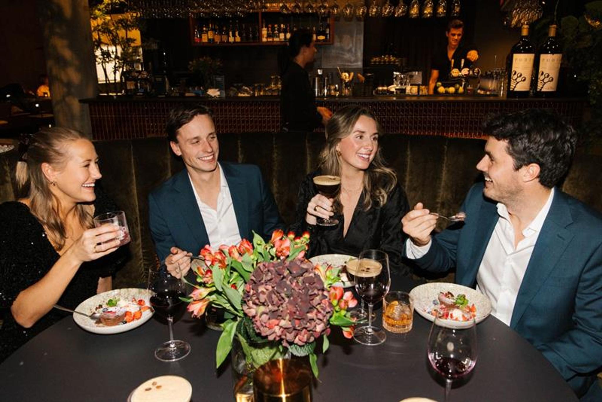En gruppe på fire personer sitter rundt et restaurantbord, smilende og pratende mens de nyter mat og drikke i et stemningsfullt miljø.