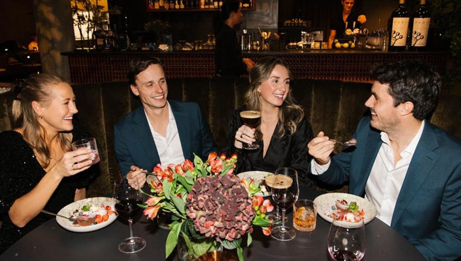En gruppe på fire personer sitter rundt et restaurantbord, smilende og pratende mens de nyter mat og drikke i et stemningsfullt miljø.