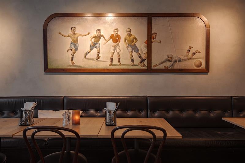 Klassisk restaurantinteriør med polstrede benker, trebord og et veggmaleri av fotballspillere.