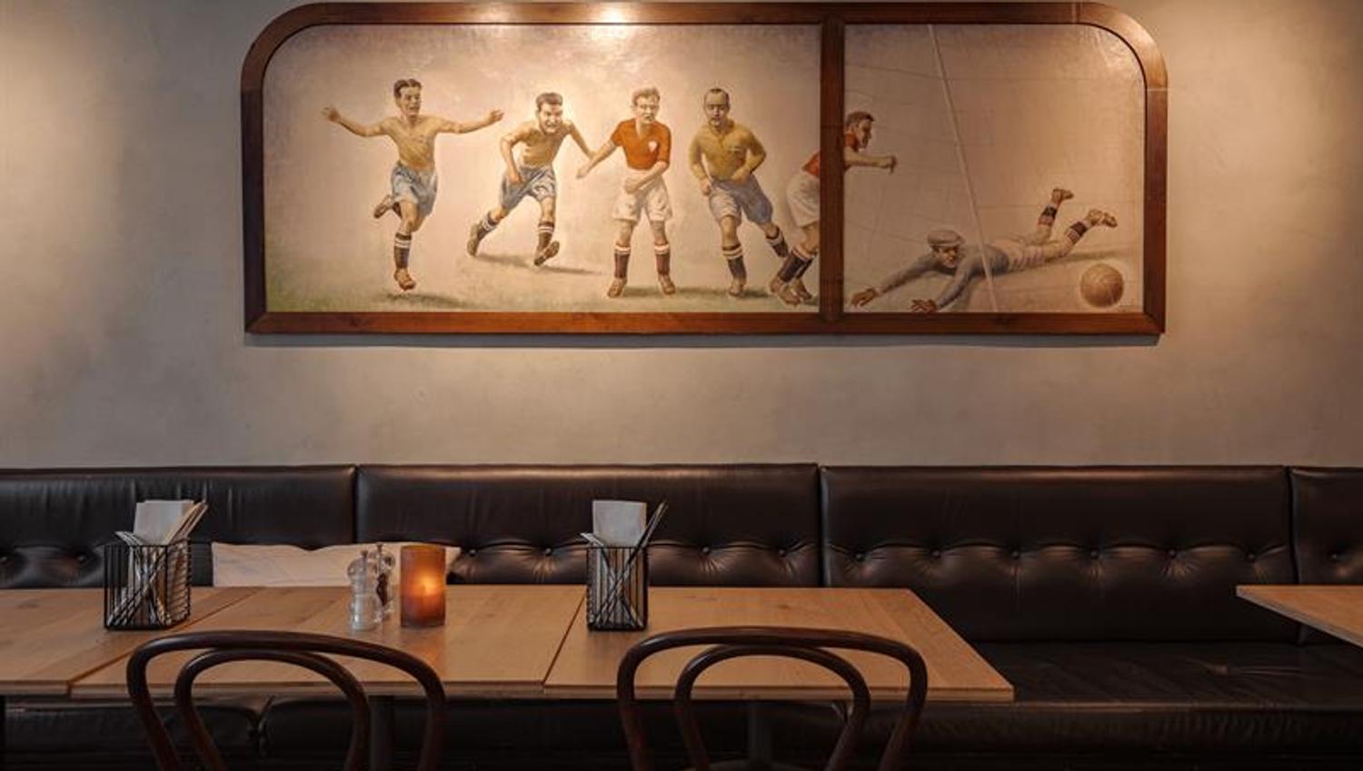 Klassisk restaurantinteriør med polstrede benker, trebord og et veggmaleri av fotballspillere.