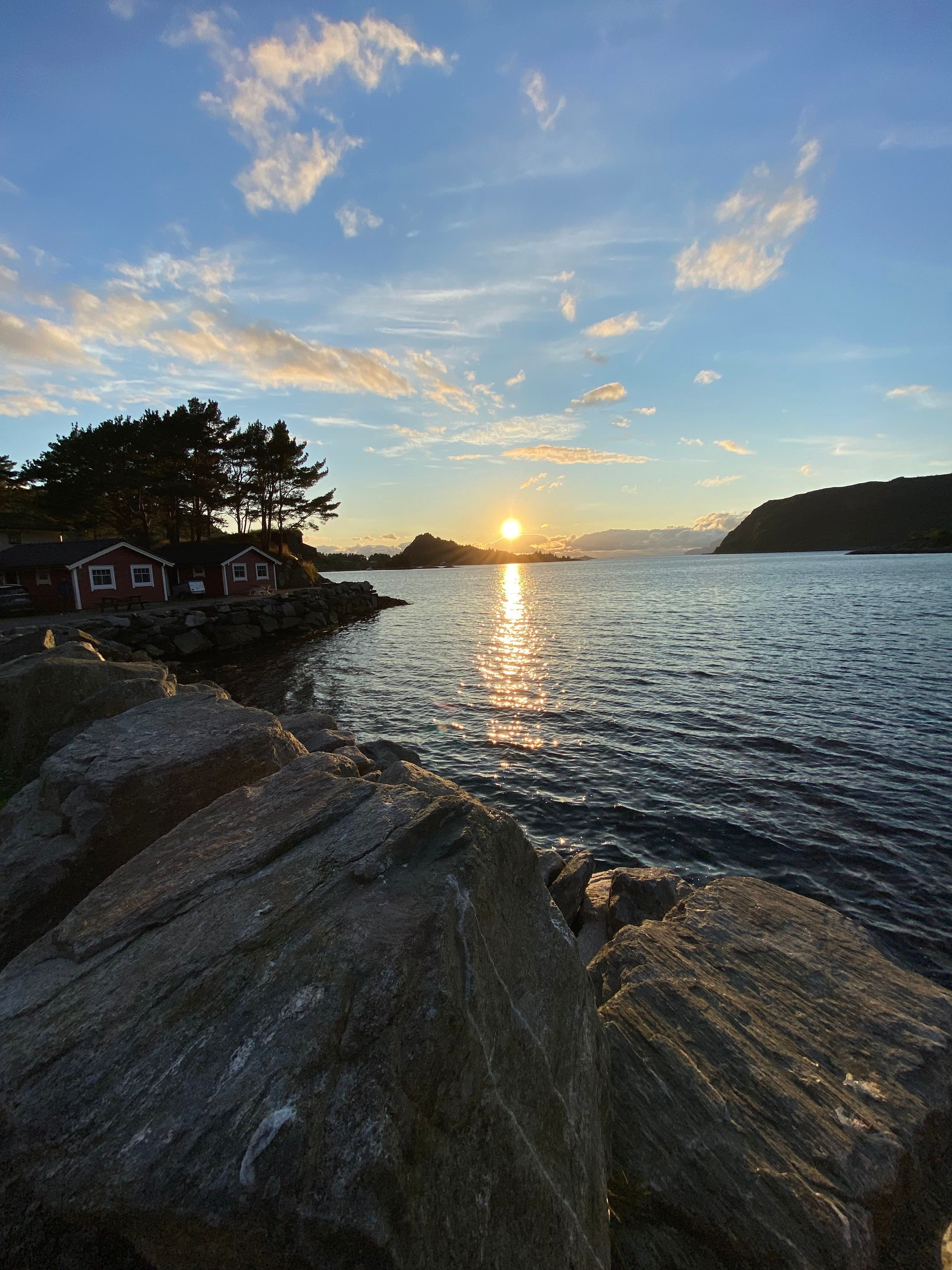 Low sun sets over a calm bay, reflecting on the water beside rocky shore and small cabins under scattered clouds..