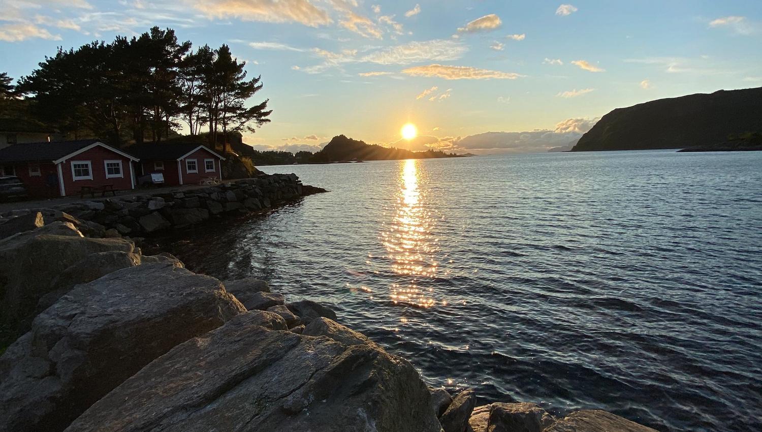 Low sun sets over a calm bay, reflecting on the water beside rocky shore and small cabins under scattered clouds..