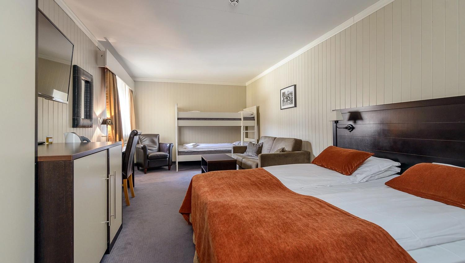 Romslig hotellrom med dobbeltseng i forgrunnen, køyeseng bakerst, sofa, lenestol, skrivebord og TV ved vindu..