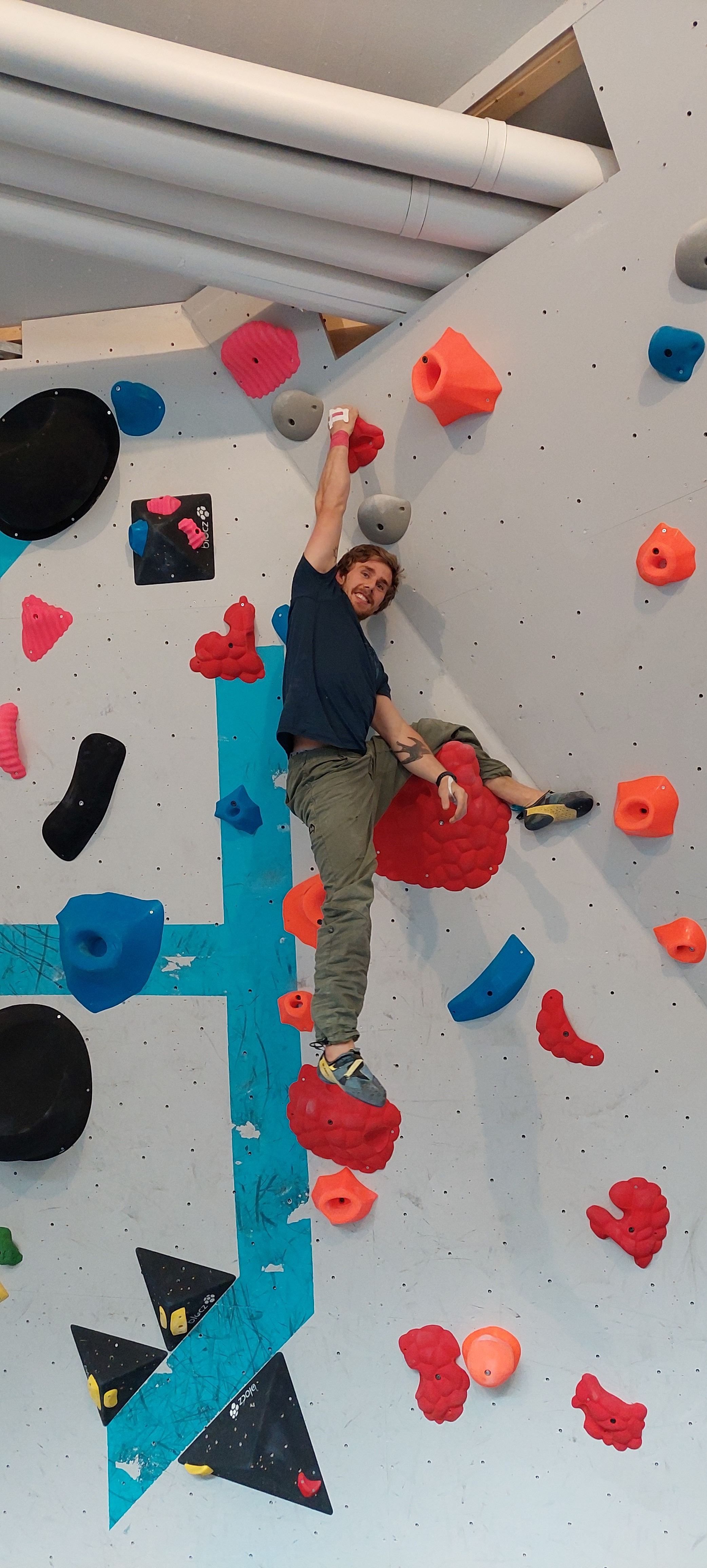 Person climbing an indoor bouldering wall, reaching up with one hand on a red hold among colorful grips..