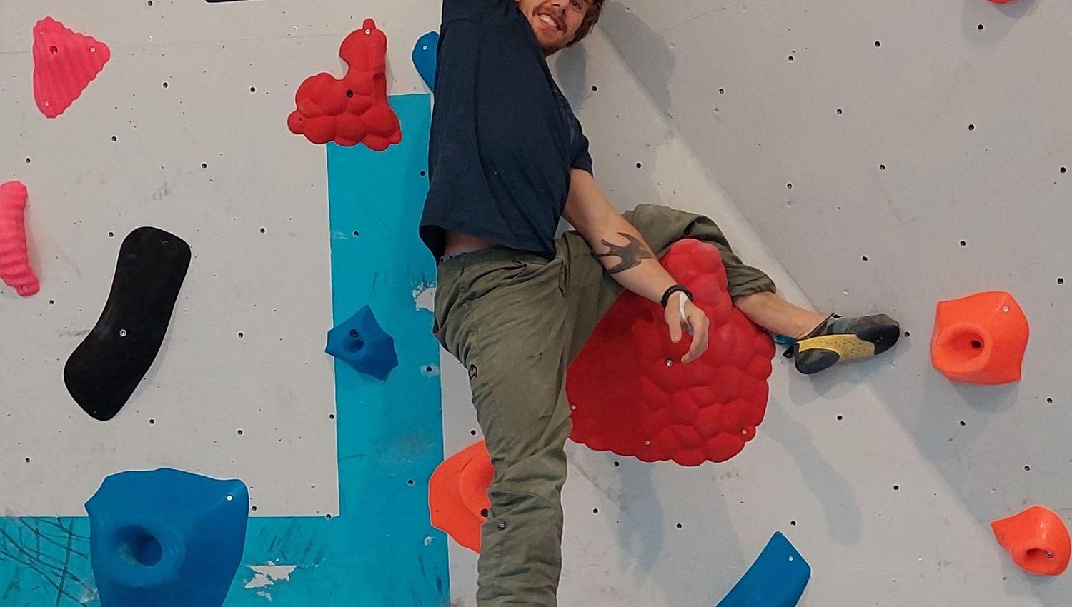 Person climbing an indoor bouldering wall, reaching up with one hand on a red hold among colorful grips..