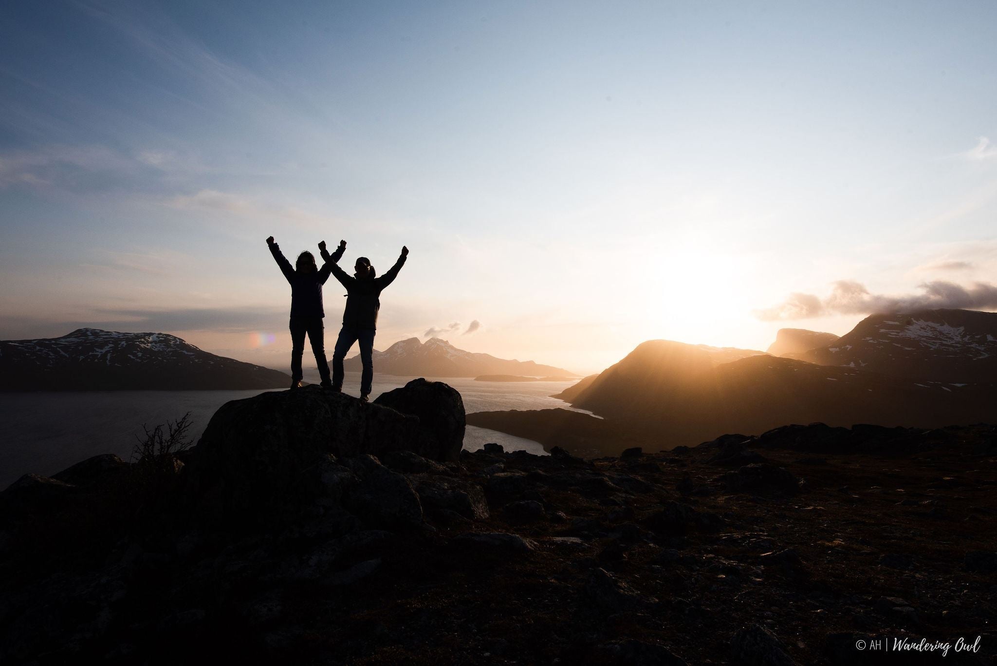 Two people raising their hands in the air in front of the midnight sun