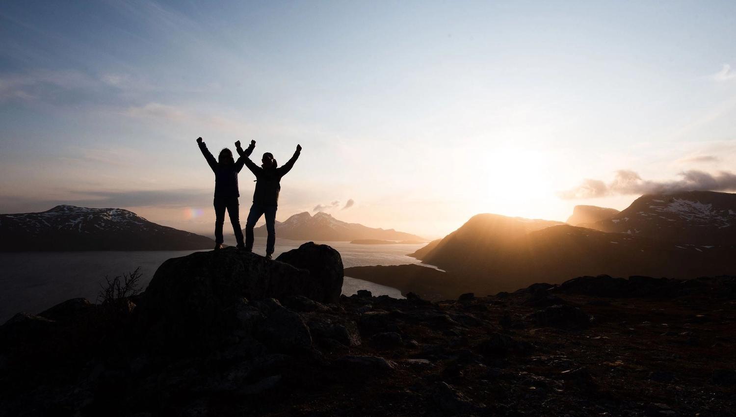 Two people raising their hands in the air in front of the midnight sun