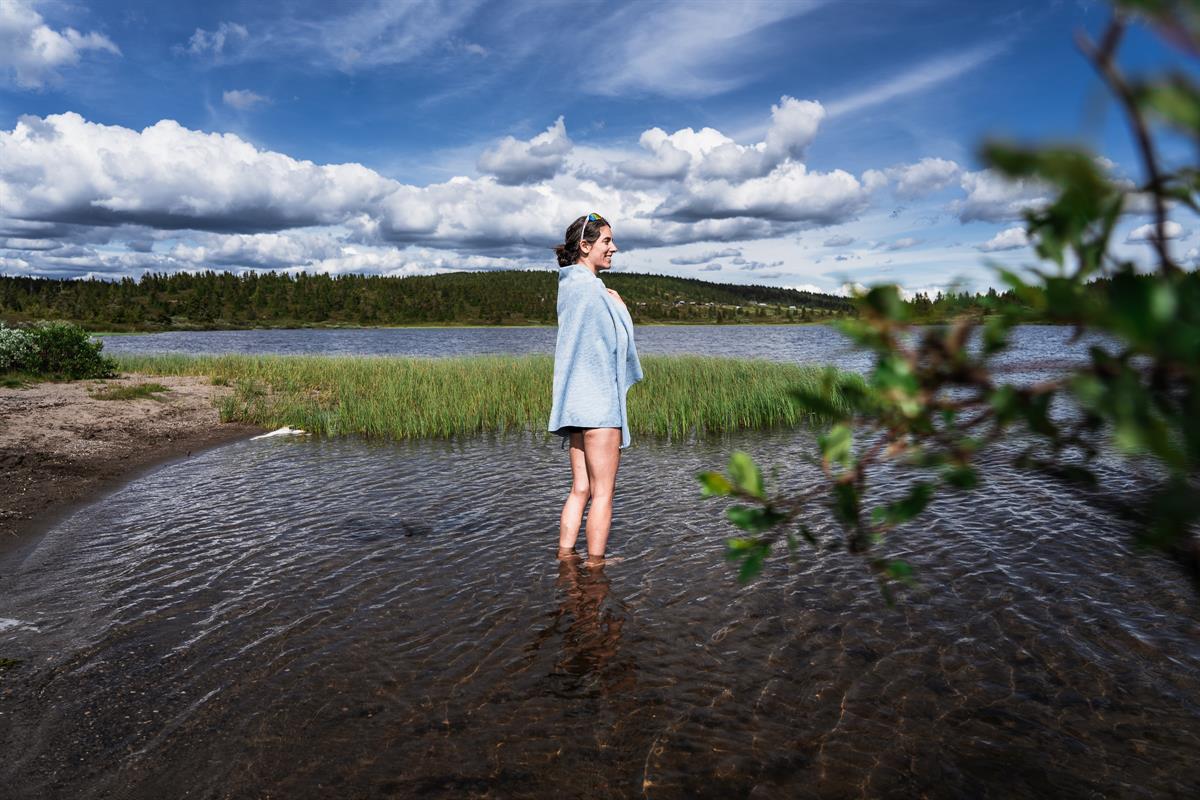 Eine Frau, in ein Handtuch gehüllt, steht im flachen Wasser eines Sees und blickt zur Seite, dahinter Schilf und Wald.