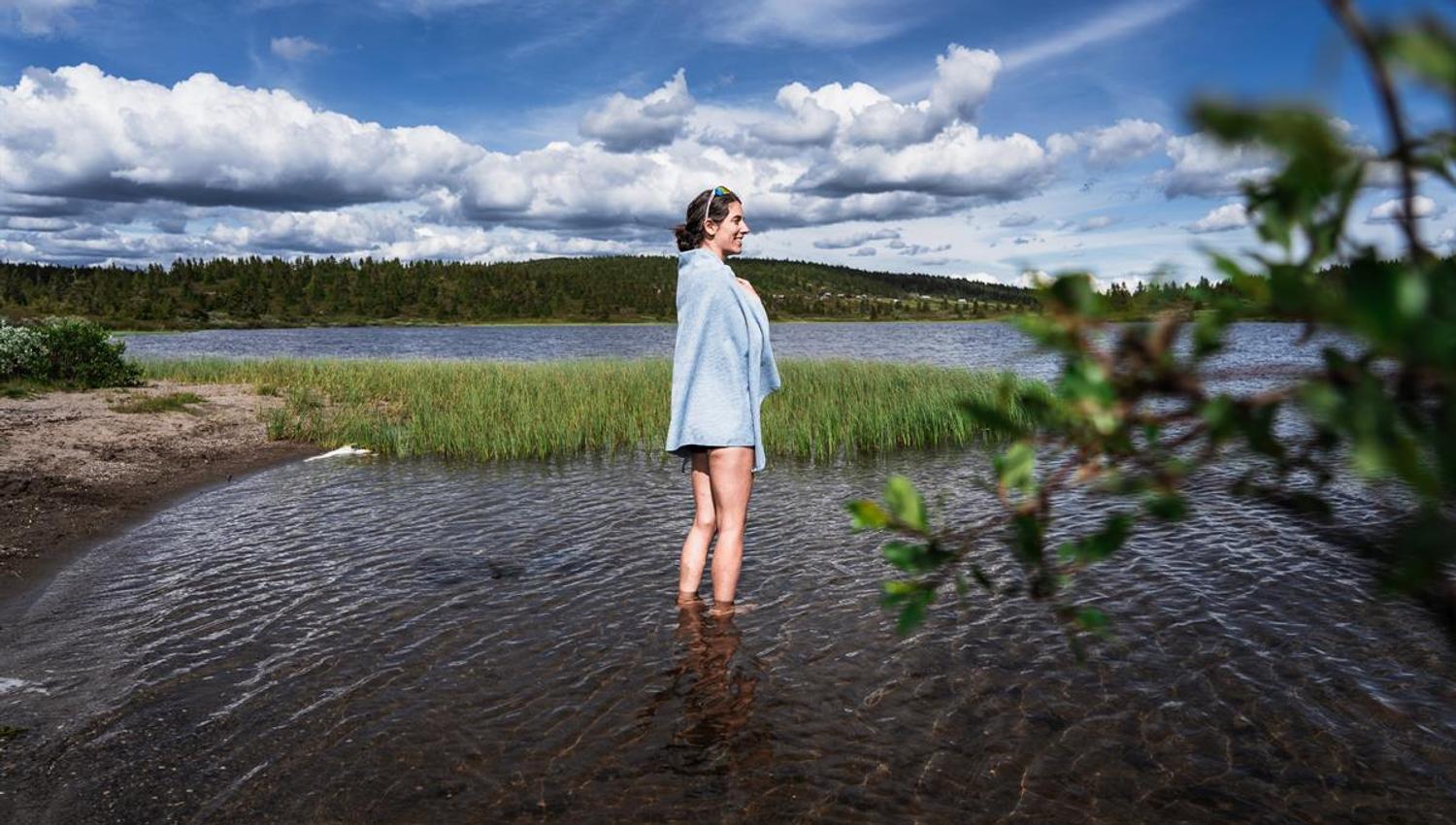 Eine Frau, in ein Handtuch gehüllt, steht im flachen Wasser eines Sees und blickt zur Seite, dahinter Schilf und Wald.