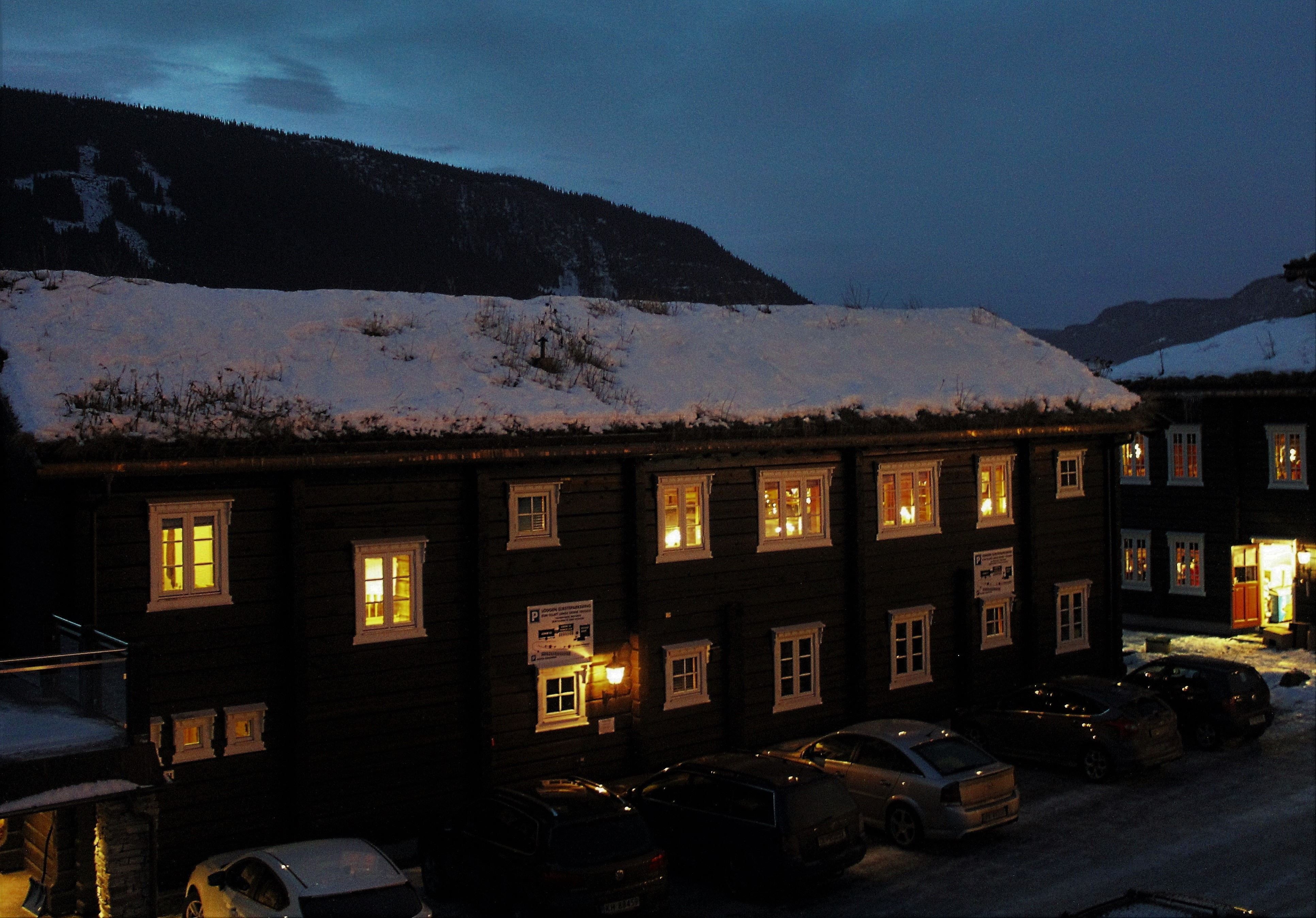 Mörk träbyggnad med snötäckt tak och upplysta fönster i vinterkväll, bilar parkerade nedanför och berg i bakgrunden.