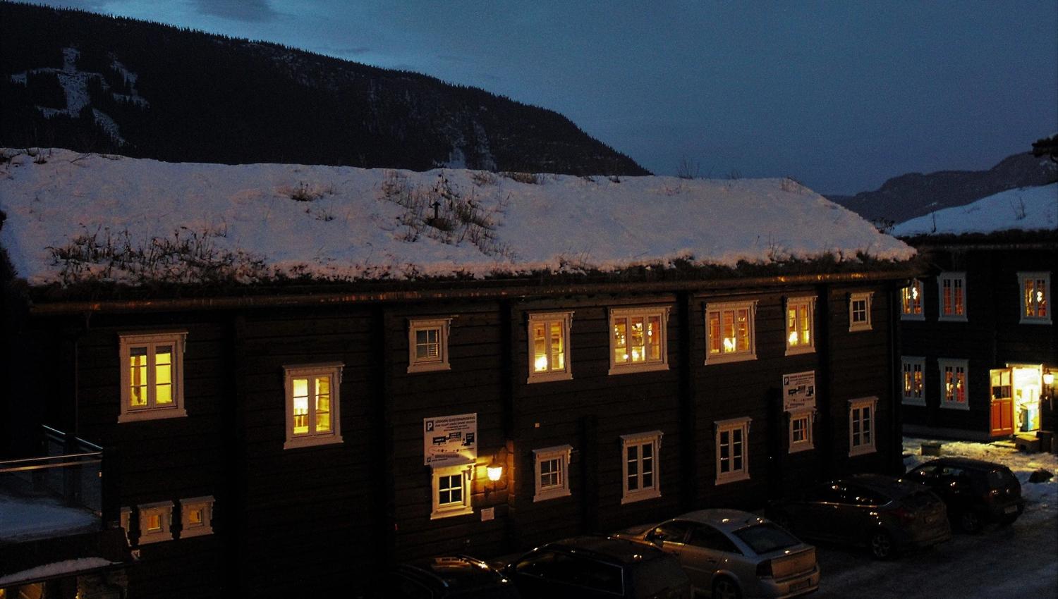 Mörk träbyggnad med snötäckt tak och upplysta fönster i vinterkväll, bilar parkerade nedanför och berg i bakgrunden.