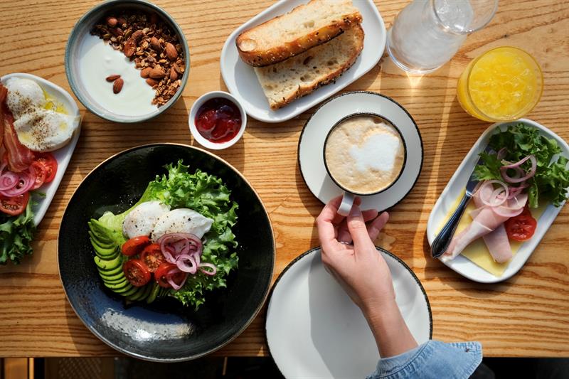 A breakfast spread on a wooden table featuring poached eggs with avocado, tomatoes, and pickled onions on a bed of lettuce, crispy bacon, granola with