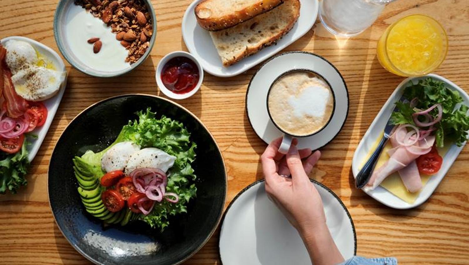 A breakfast spread on a wooden table featuring poached eggs with avocado, tomatoes, and pickled onions on a bed of lettuce, crispy bacon, granola with